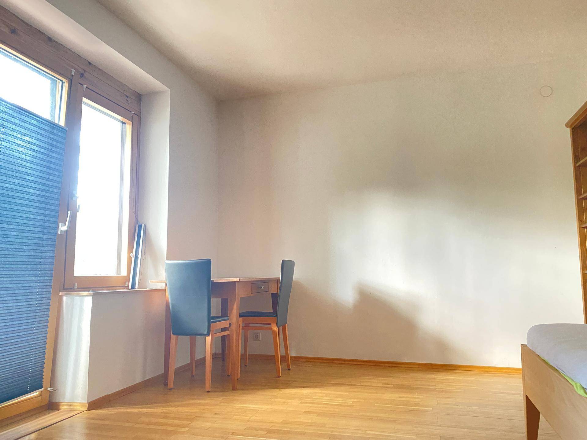 Heller Raum mit Holzboden, weiß gestrichenen Wänden und einem kleinen Essbereich am Fenster.
