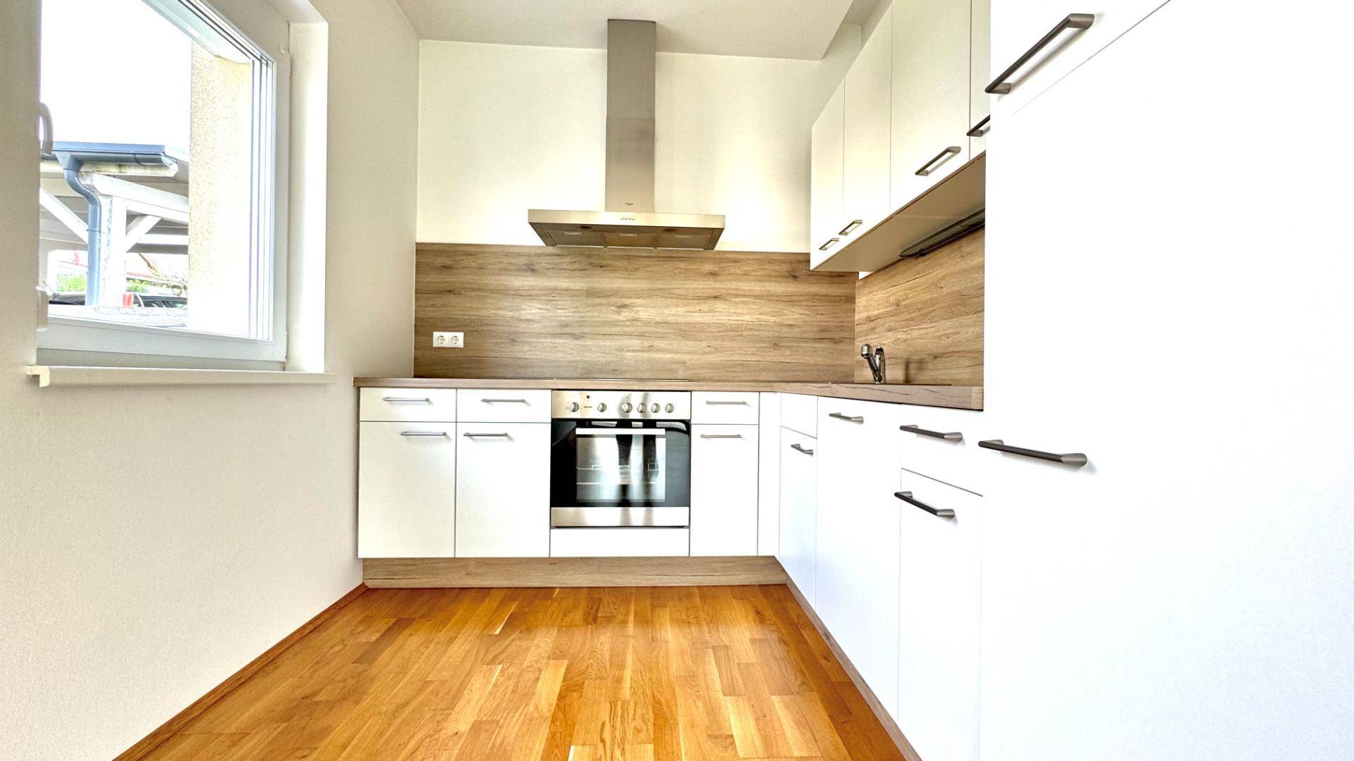 Kompakte Küchenzeile mit weißen Fronten, Holzrückwand und Fenster für Tageslicht.