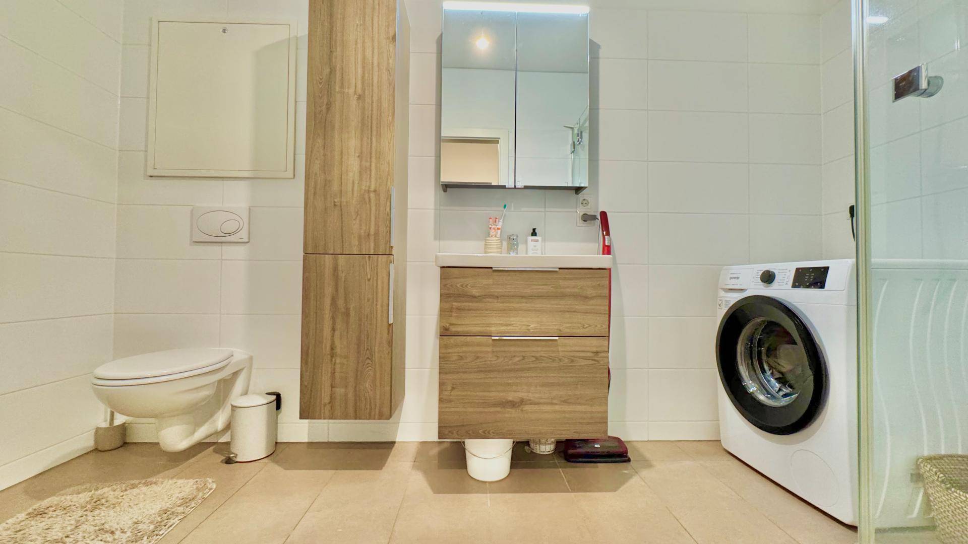 Modernes Badezimmer mit WC, Waschbecken, Dusche und integrierter Waschmaschine, hell gefliest.