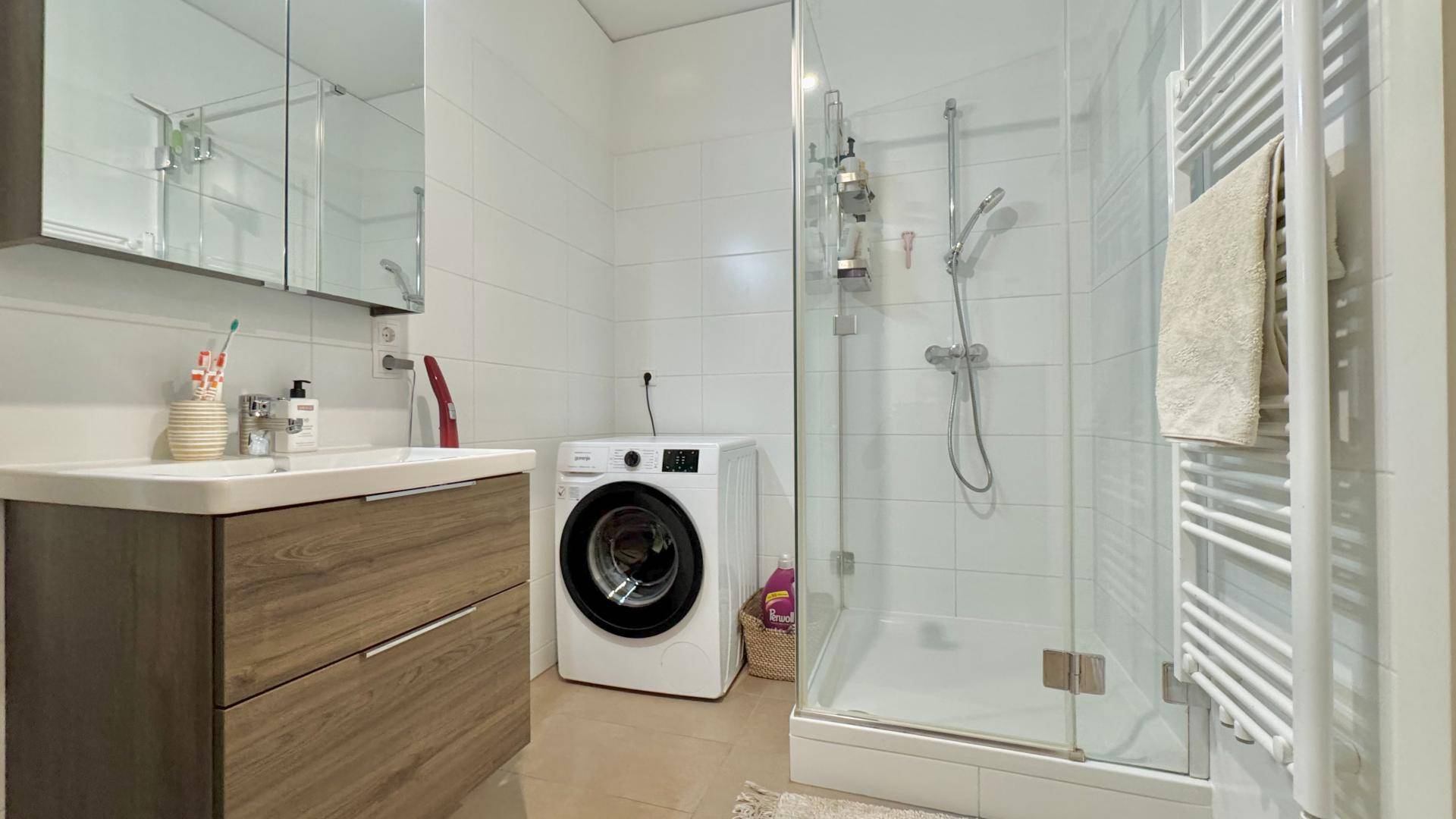 Funktionales Badezimmer mit ebenerdiger Dusche, Waschbecken, Waschmaschine und Handtuchheizkörper.