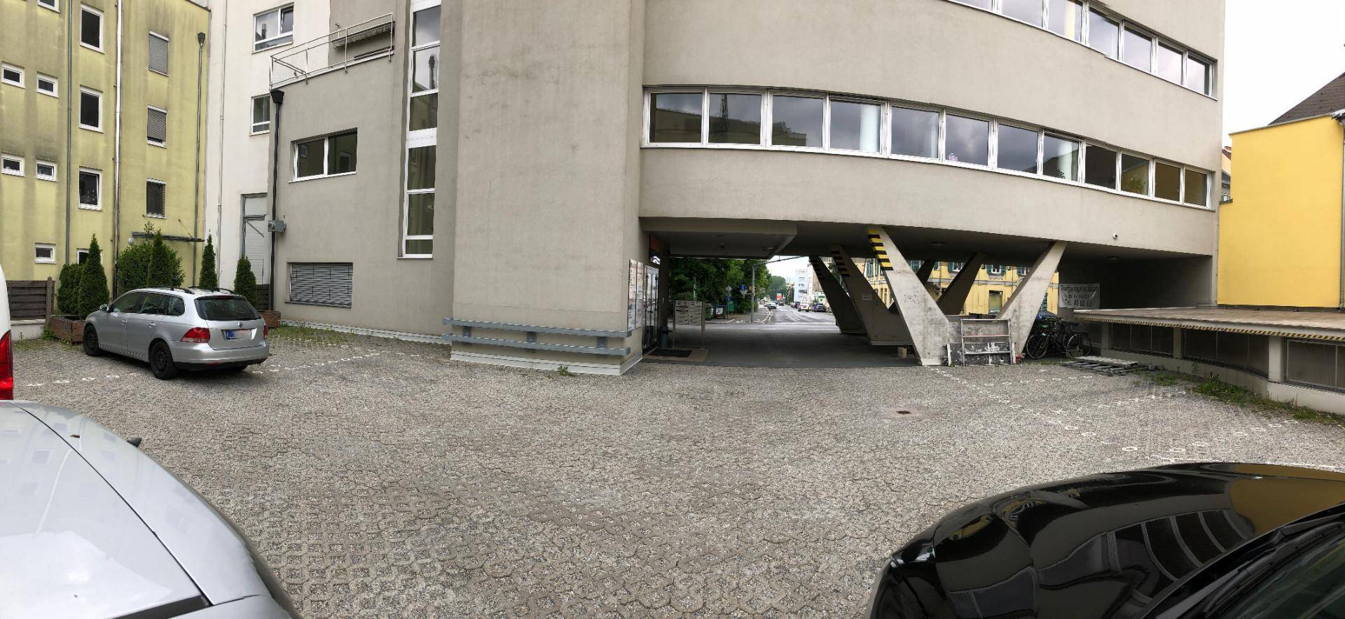 Rückseite eines Gebäudes mit gepflastertem Parkplatz und Zugang zu einem überdachten Bereich.