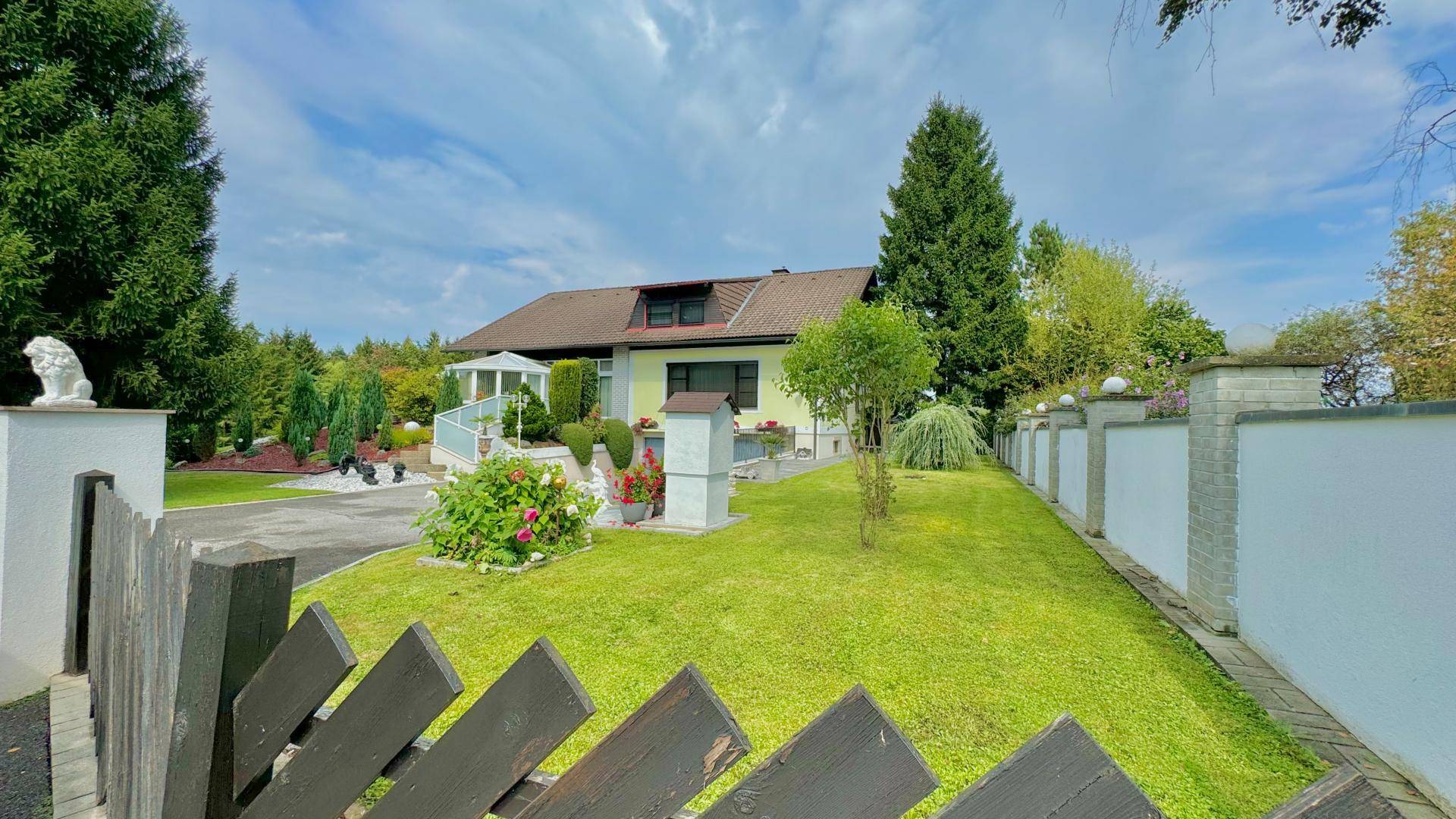 Vorgarten mit Zaun, Rasenfläche und dem Haus im Hintergrund unter blauem Himmel.
