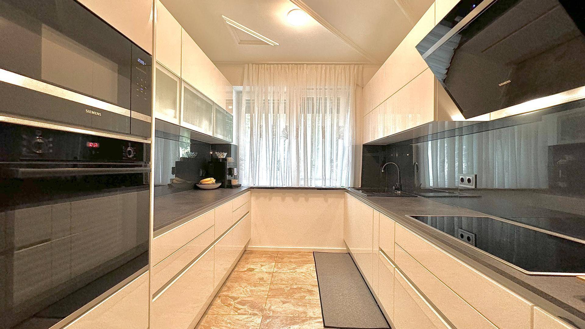 Moderne Küche mit weißen Hochglanzfronten, Einbaugeräten und Fenster mit Vorhängen.