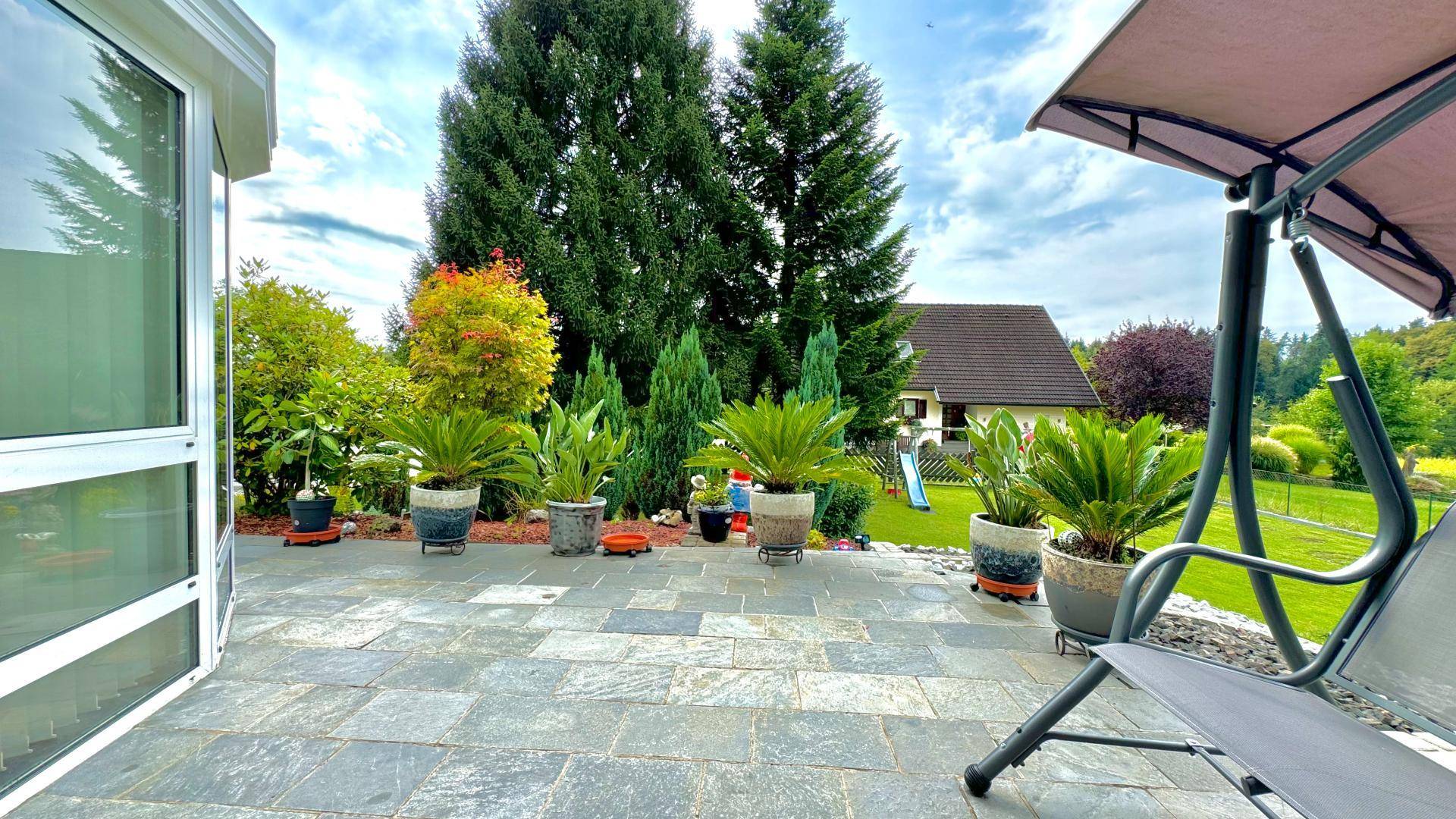 Große gepflasterte Terrasse mit Topfpflanzen und einer Gartenschaukel, umgeben von Bäumen.