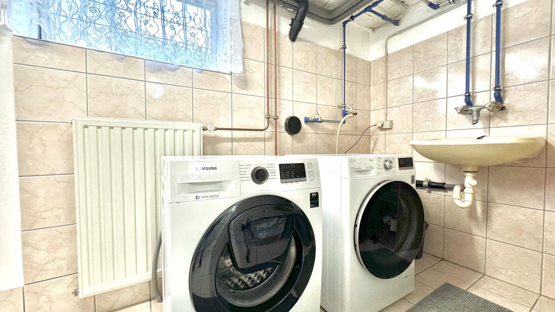Waschküche mit zwei Waschmaschinen, Waschbecken und sichtbaren Installationen an der Wand.