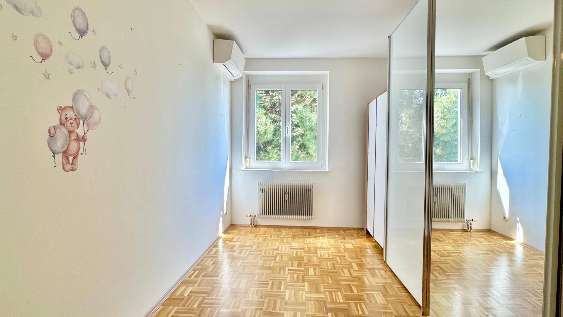 Kinderzimmer mit Parkettboden, Klimaanlage und Fenster mit Blick ins Grüne.