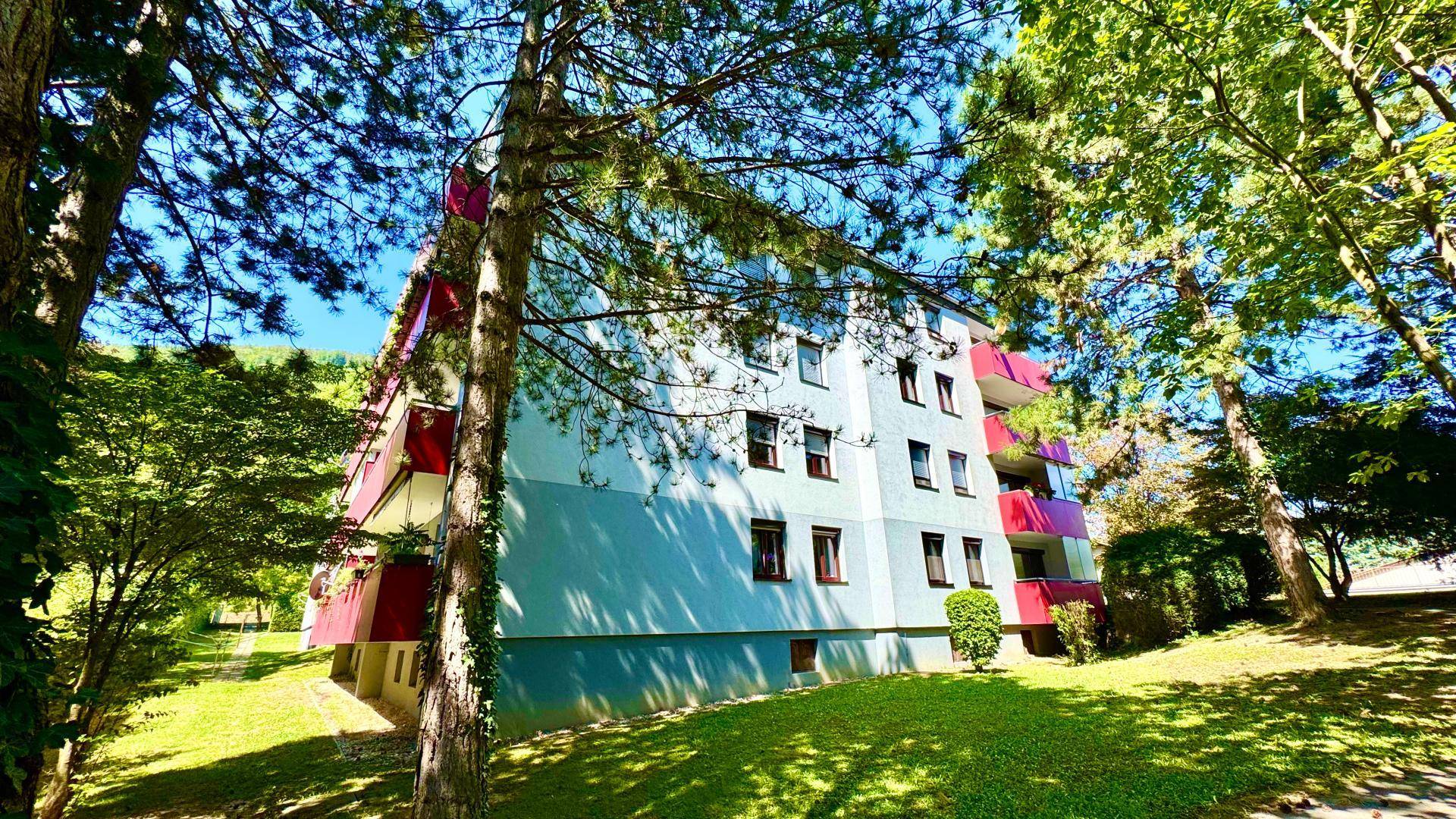 Mehrfamilienhaus umgeben von Bäumen und Grünflächen unter strahlend blauem Himmel.