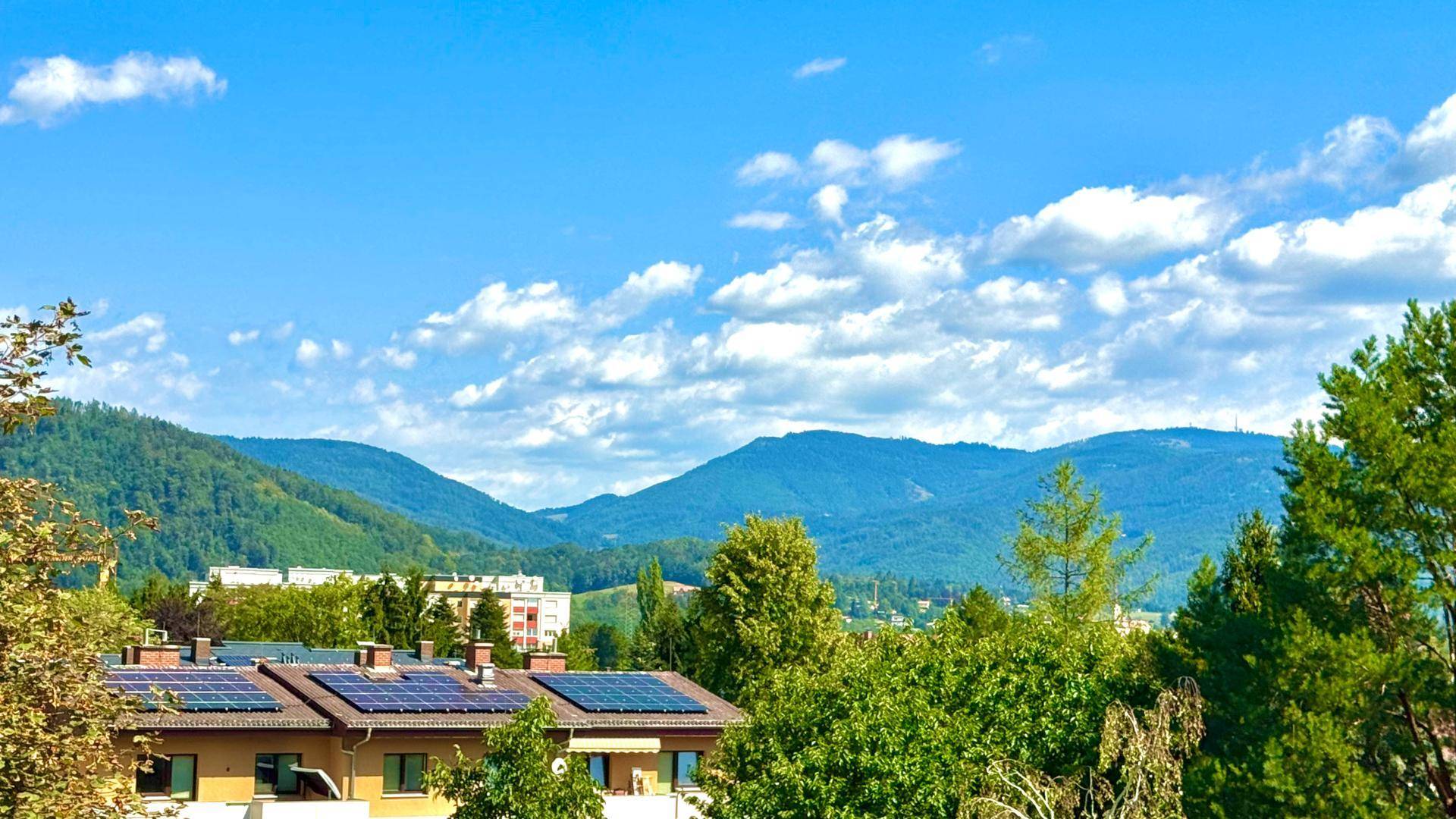 Panoramablick auf grüne Berge und Dächer mit Solaranlagen unter blauem Himmel.