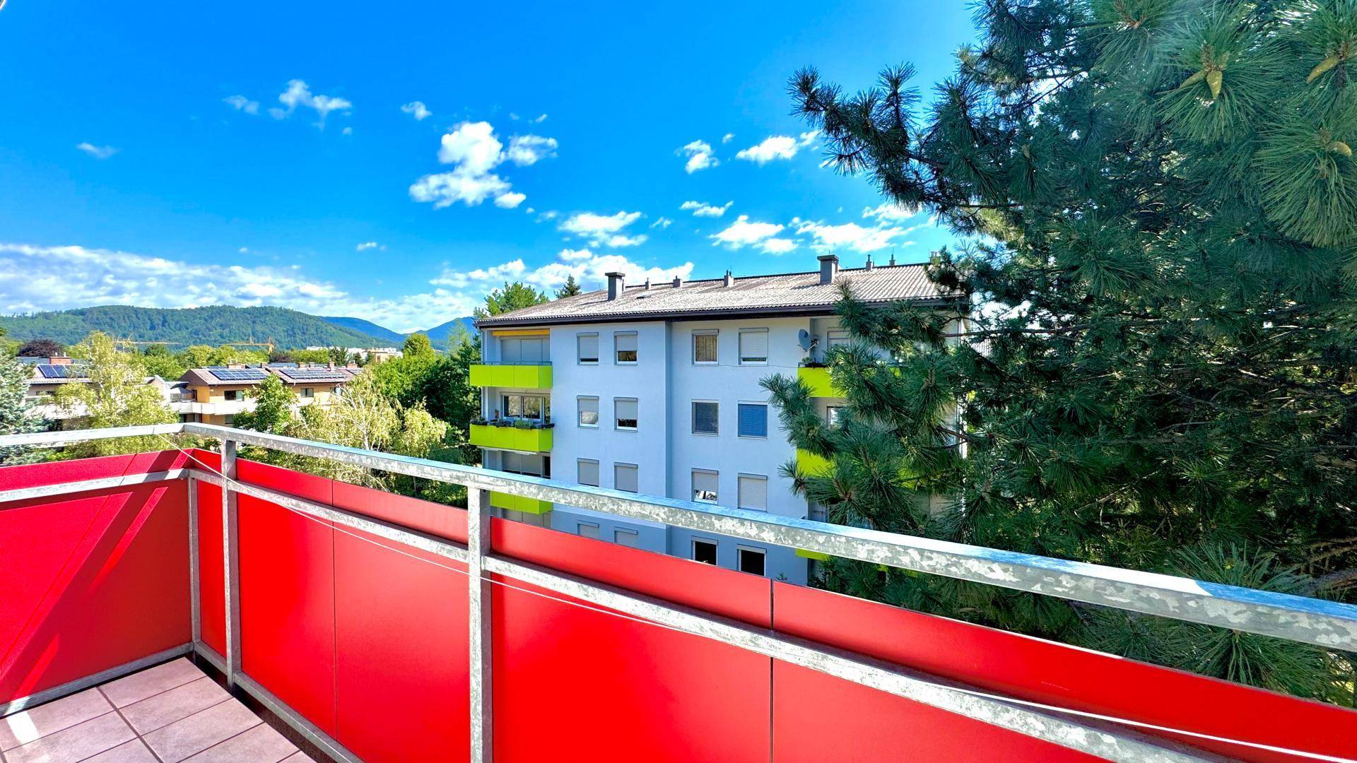 Balkon mit rotem Geländer und Blick auf die Nachbargebäude und grüne Umgebung.