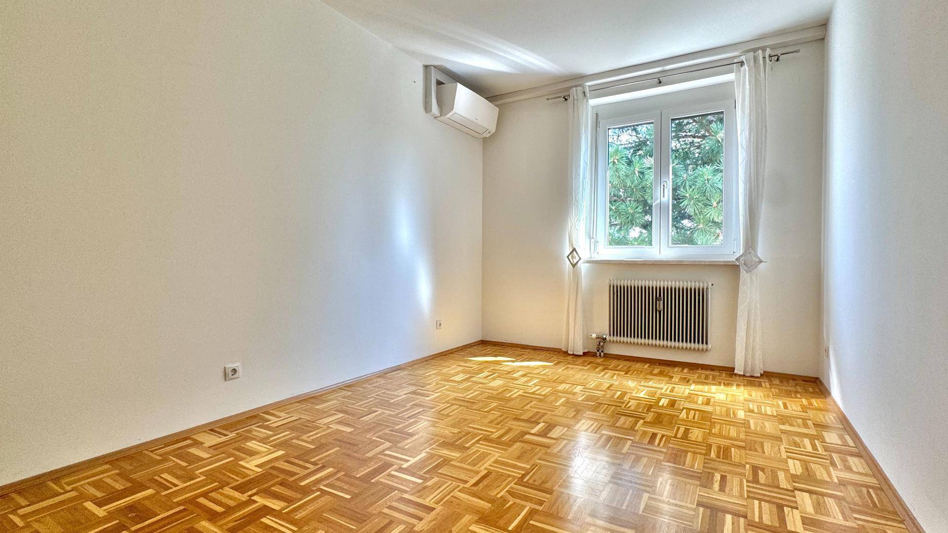 Schlafzimmer mit Parkettboden, Klimaanlage und Vorhängen am Fenster.