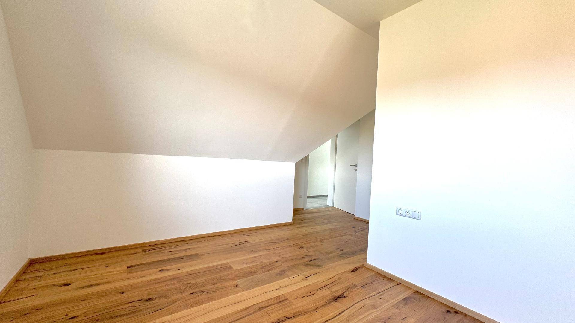 Helles, leeres Zimmer im Dachgeschoss mit Holzboden und schrägen Wänden, bereit zur Einrichtung.