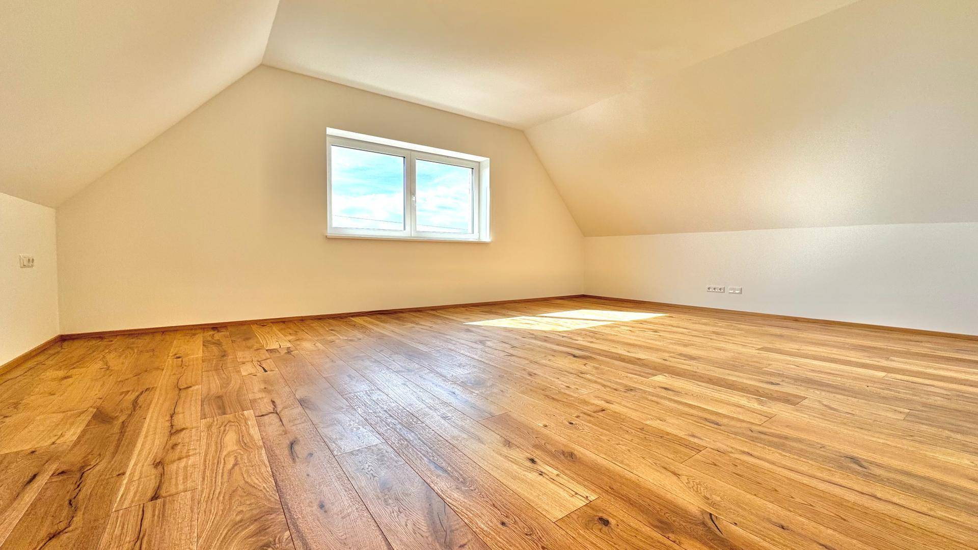Geräumiges Dachzimmer mit Holzboden, schrägen Wänden und einem Fenster für natürliche Belichtung.