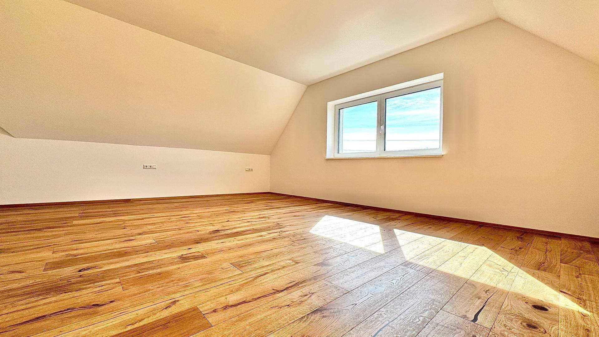 Helles Dachzimmer mit schrägen Wänden, Holzboden und einem Fenster, das viel Licht spendet.