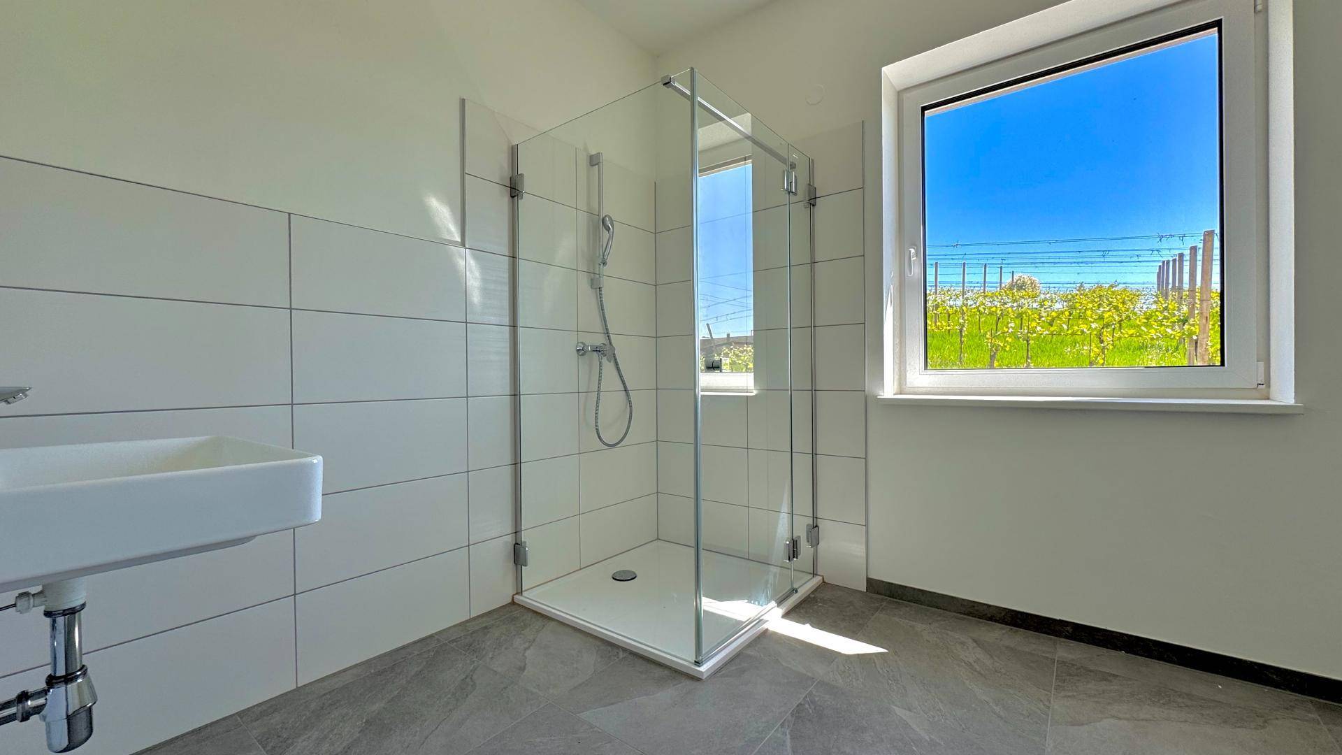 Modernes Badezimmer mit weiß gefliesten Wänden, Glasdusche und Fenster mit Blick auf die Weinberge.