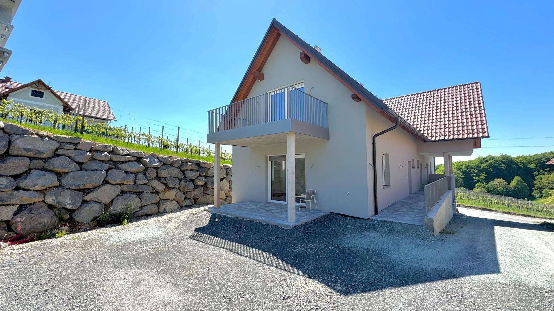Neubauhaus mit Balkon, gepflasterter Terrasse und Steinmauer, umgeben von Weinbergen.