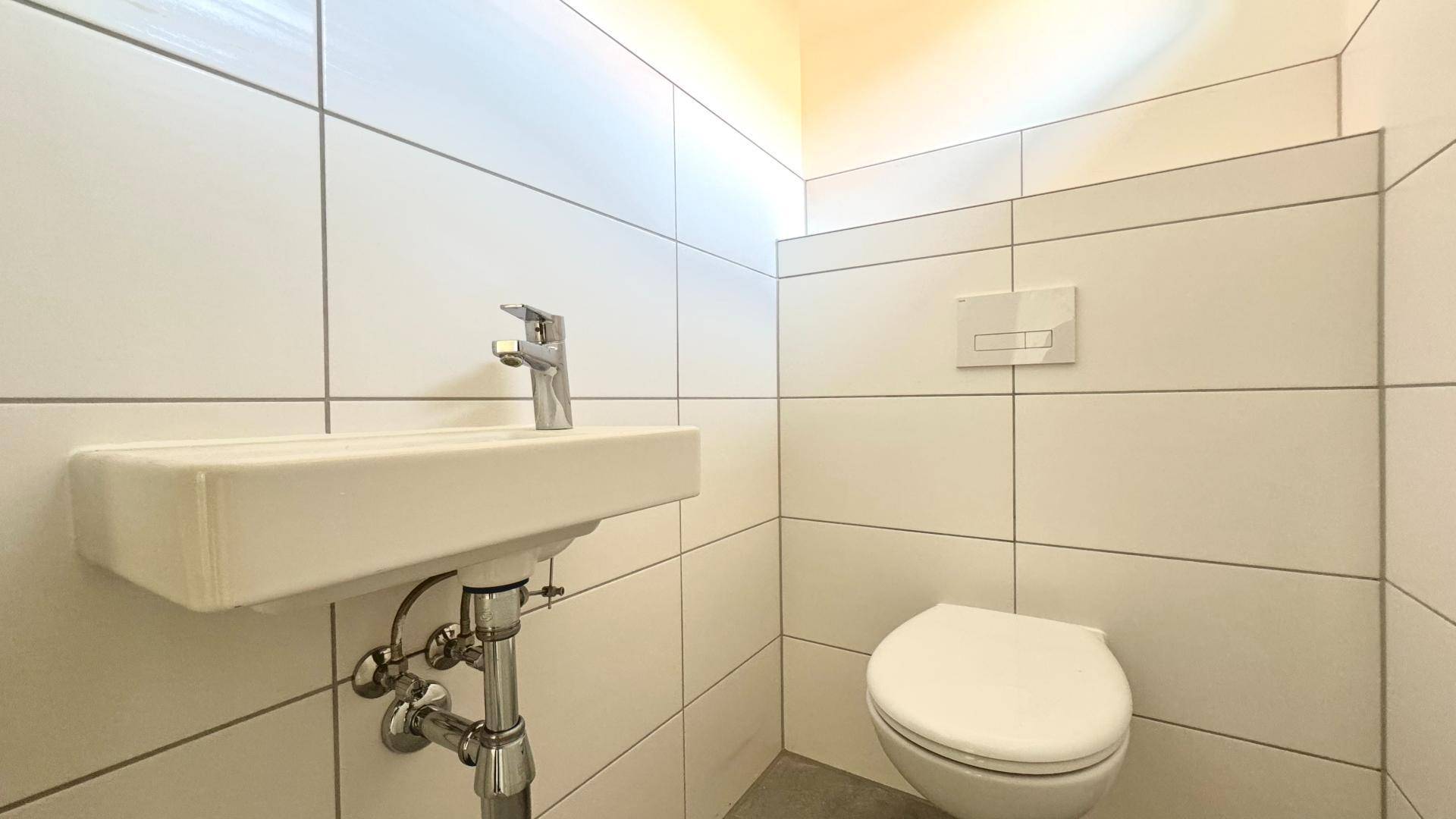 Helles Gäste-WC mit weißen Fliesen, wandhängendem WC und kleinem Handwaschbecken.