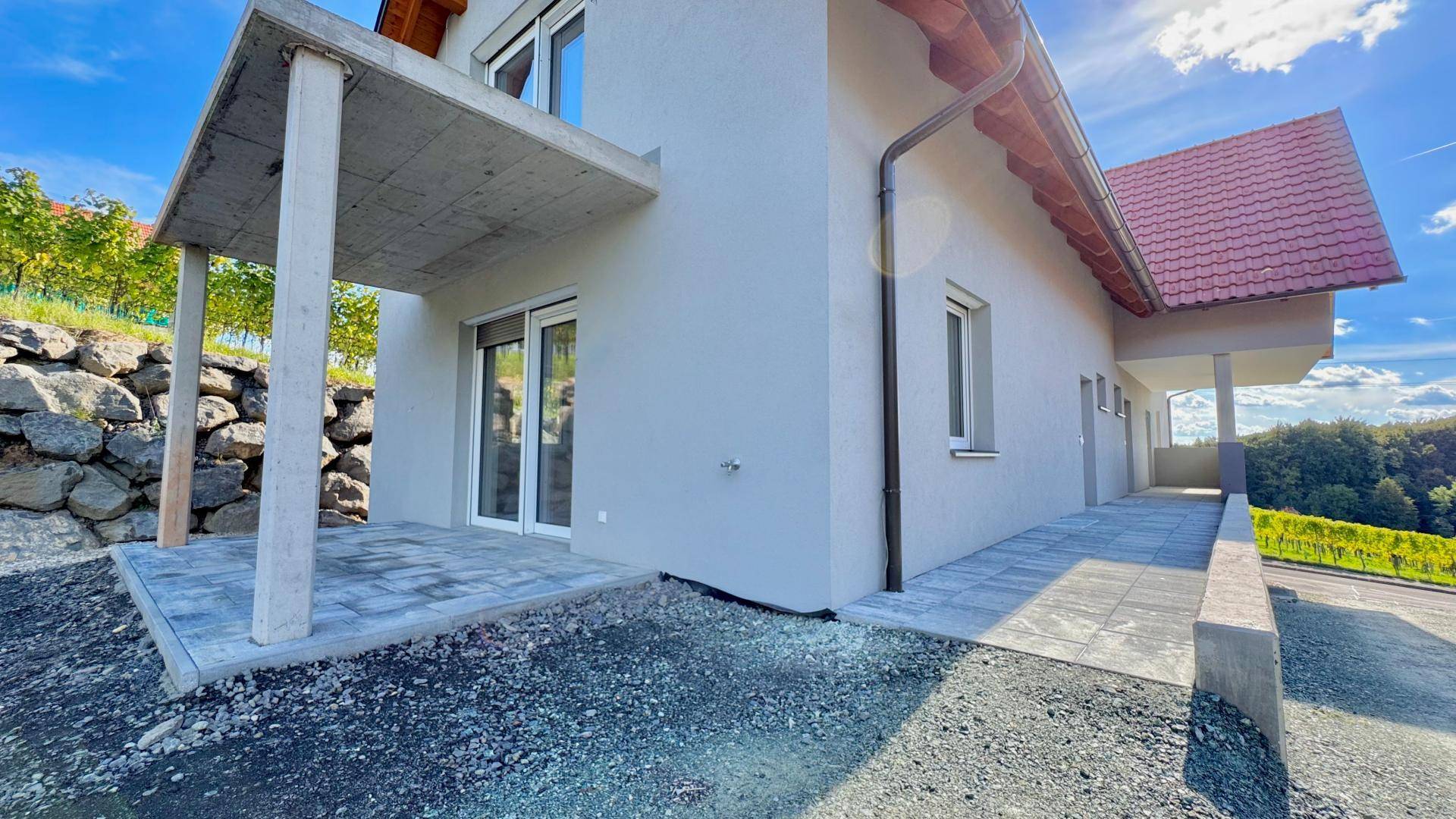 Seitenansicht des Neubaus mit gepflasterter Terrasse, Rampe und einer Natursteinmauer.