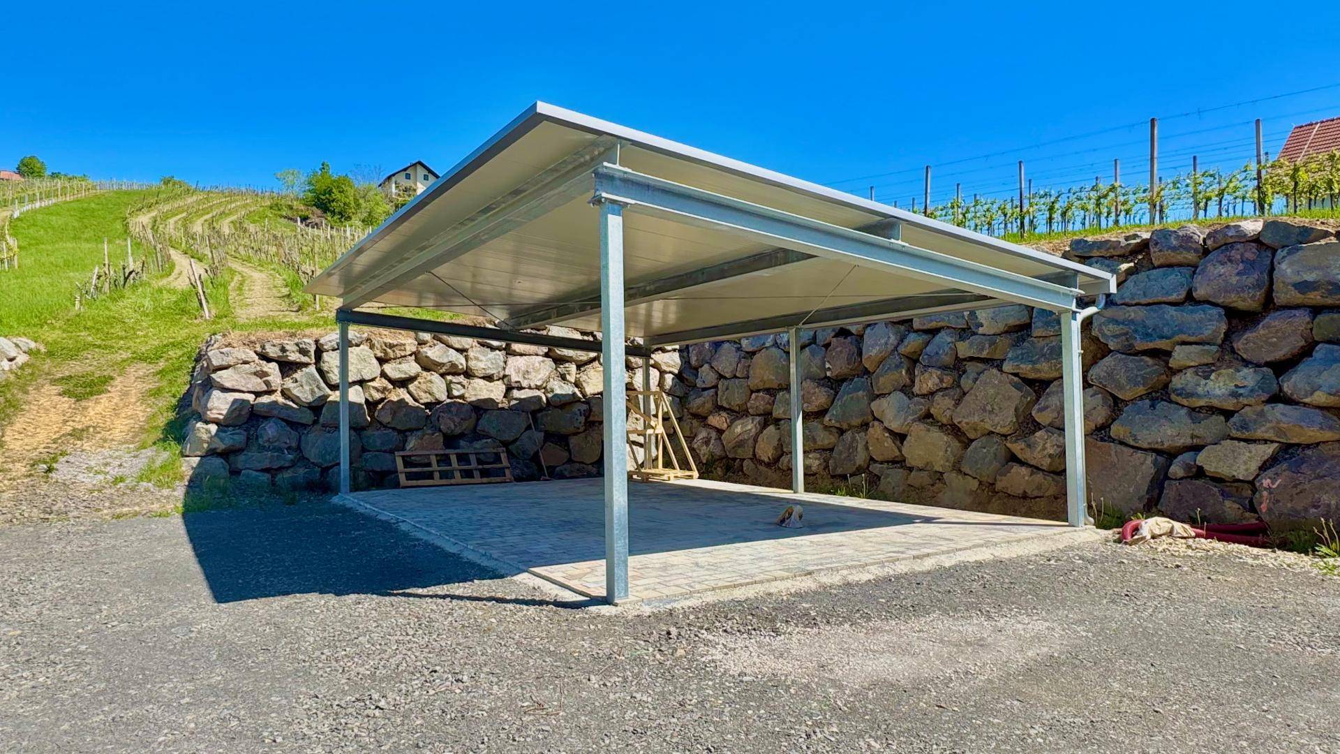 Moderner Carport mit Metallkonstruktion neben einer Steinmauer und Weinbergen.