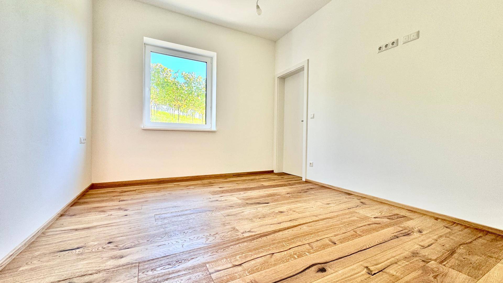Heller, leerer Raum mit Holzboden und einem Fenster mit Blick ins Grüne.