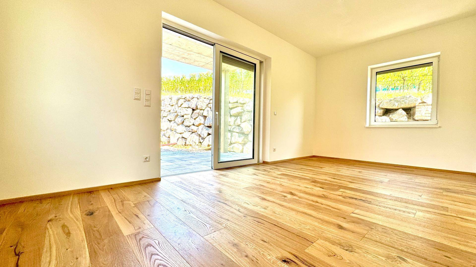 Lichtdurchfluteter Raum mit Holzboden und Zugang zu einer gepflasterten Terrasse mit Steinmauer.