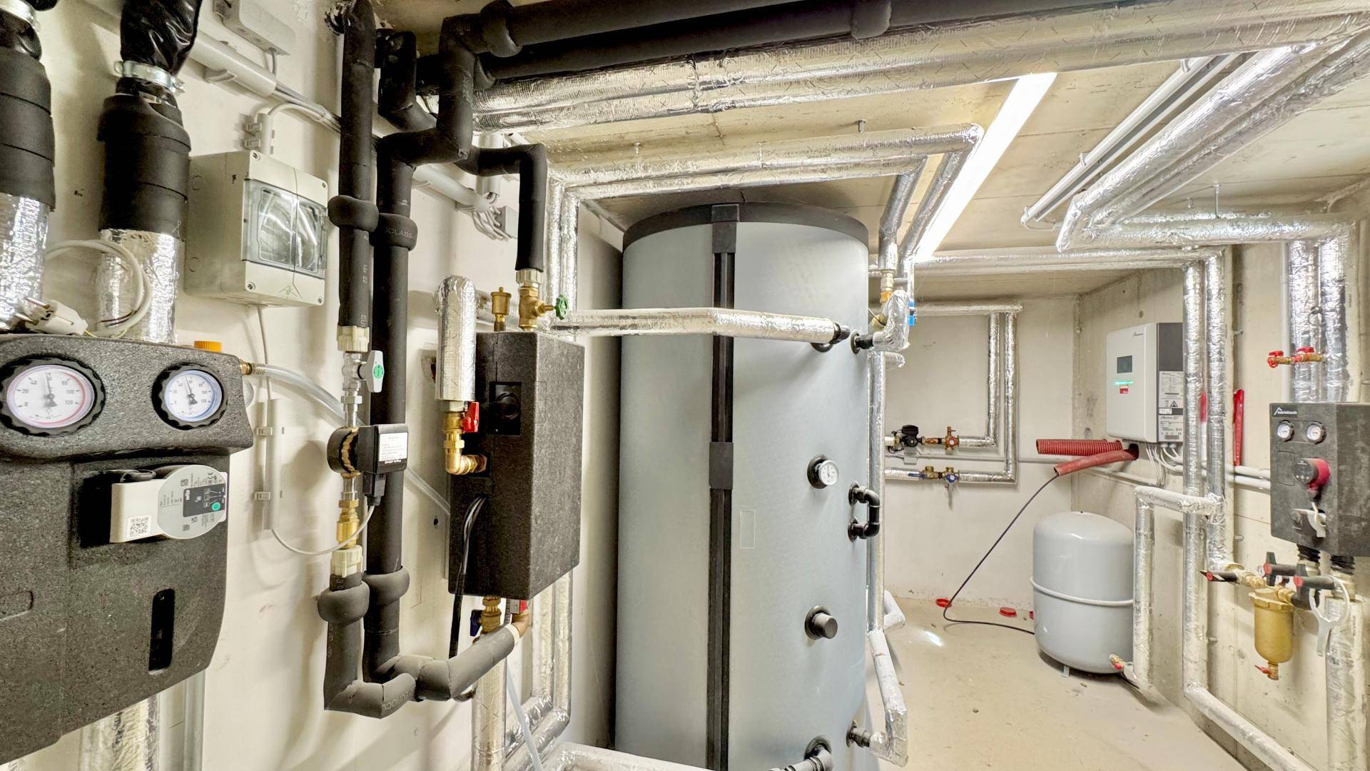 Technikraum im Keller mit Warmwasserspeicher und zahlreichen Rohrleitungen.