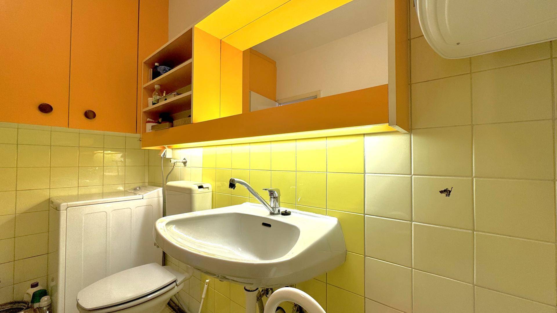 Detailansicht des Badezimmers mit gelben Fliesen, Waschbecken und einem orangefarbenen Spiegelschrank.