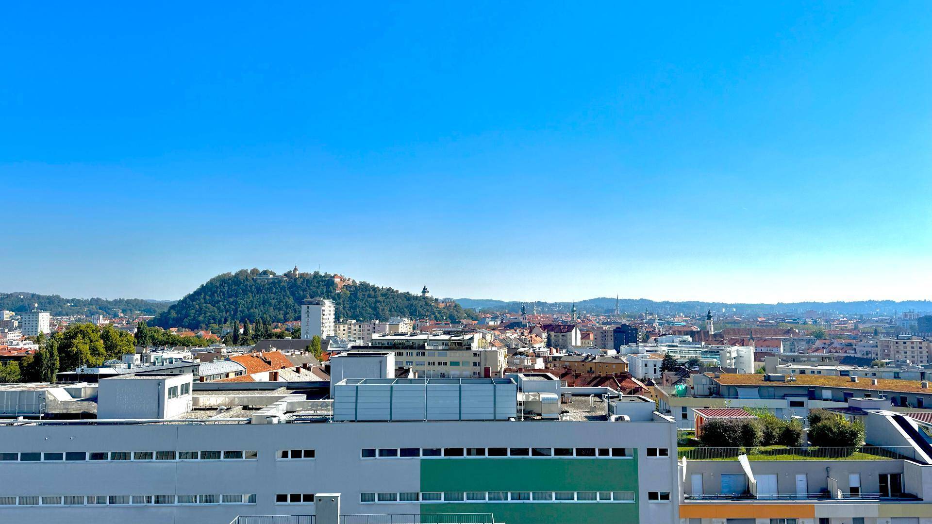 Panoramablick über die Stadt mit grünen Hügeln und urbaner Bebauung unter blauem Himmel.
