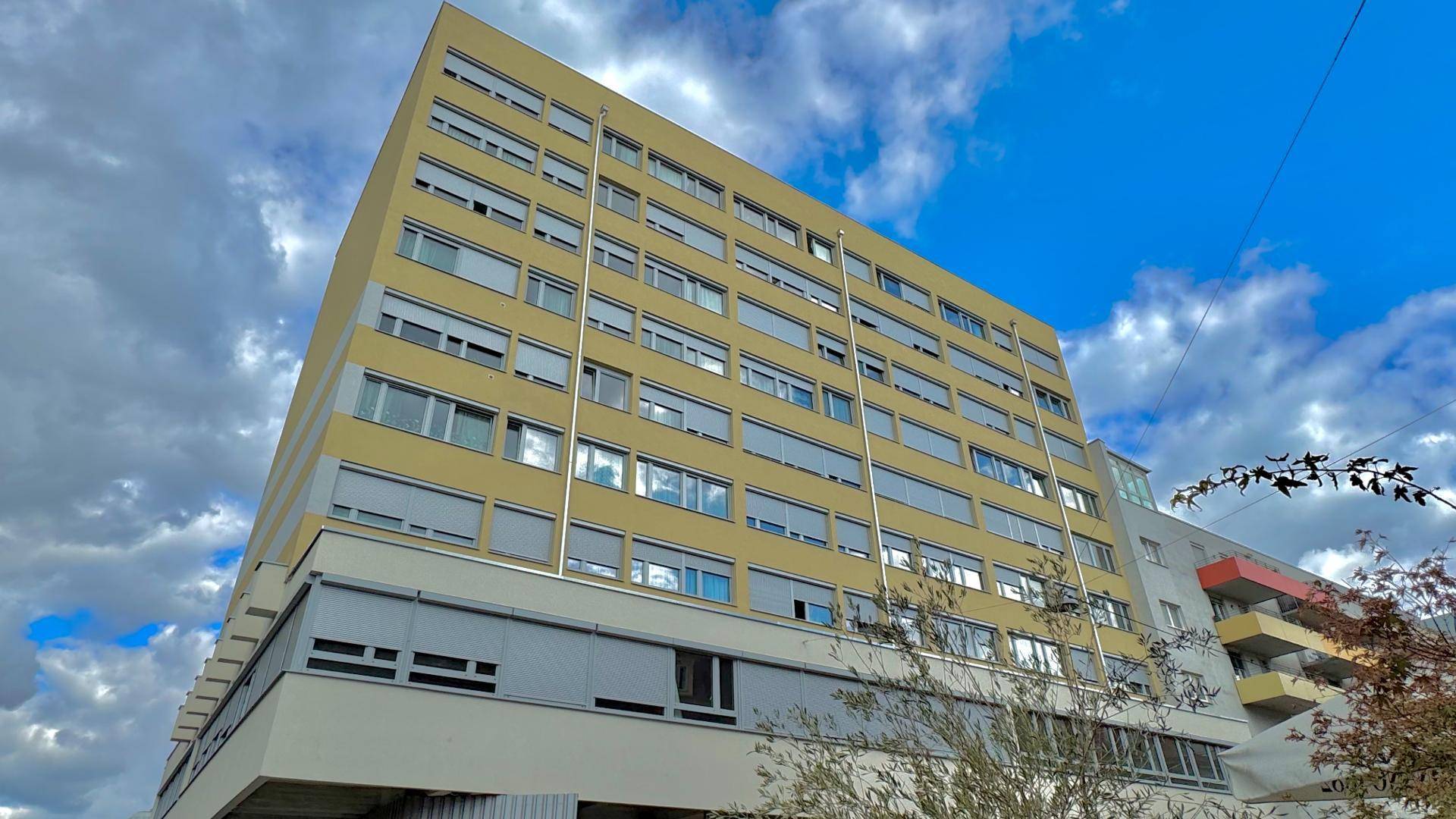 Mehrstöckiges Wohngebäude mit gelber Fassade und zahlreichen Fenstern unter bewölktem Himmel.