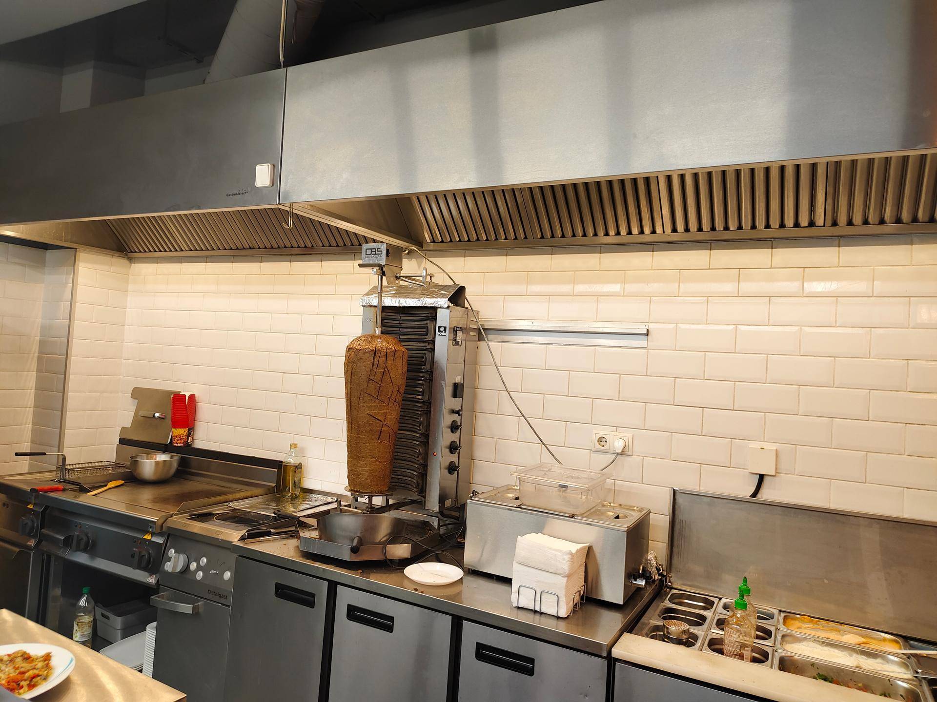 Gewerbeküche mit großem Döner-Spieß, professioneller Dunstabzugshaube und Edelstahl-Arbeitsflächen.