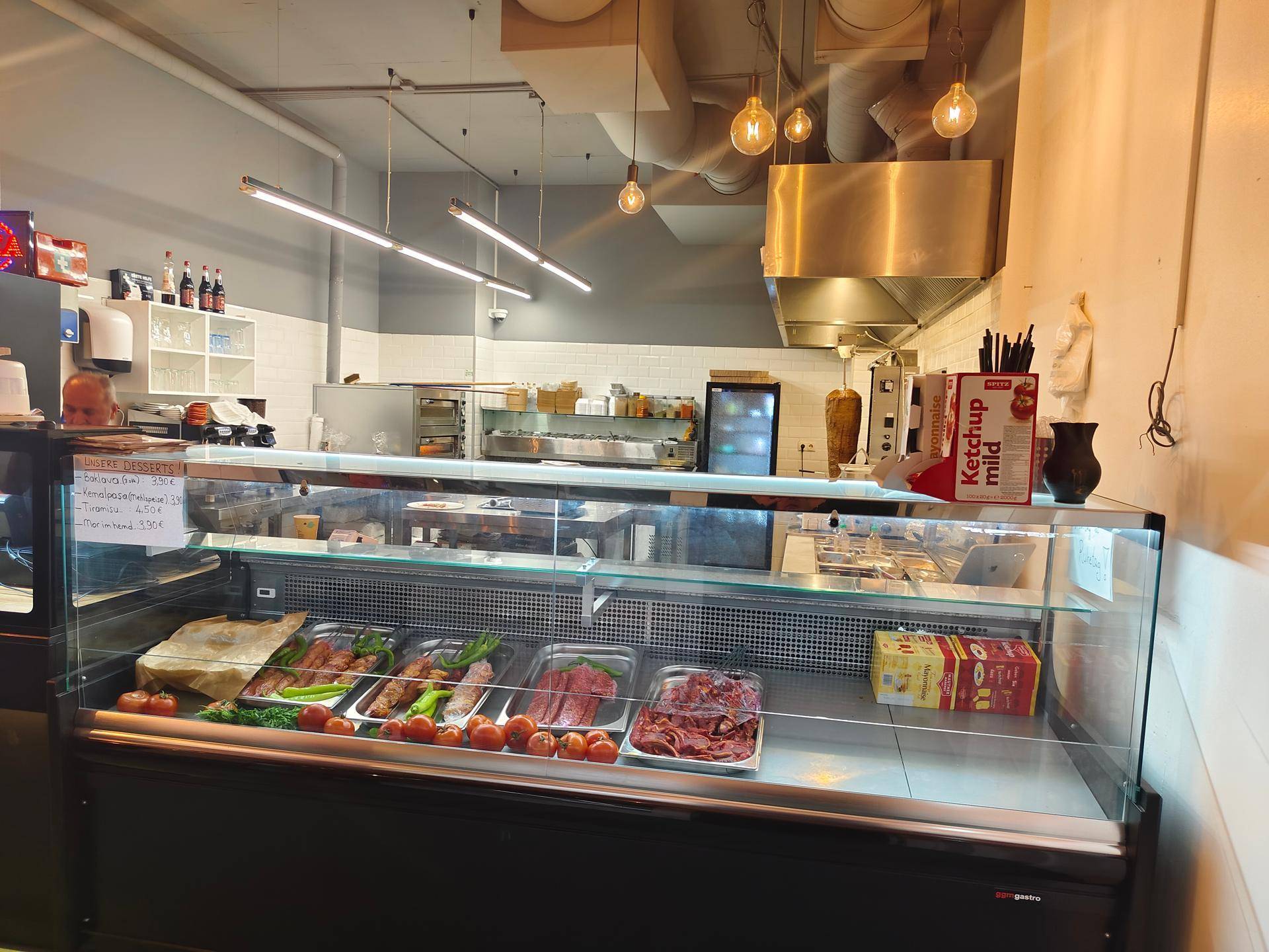 Thekenbereich mit Kühlvitrine für Fleisch und Gemüse, dahinter die professionelle Küchenausstattung.