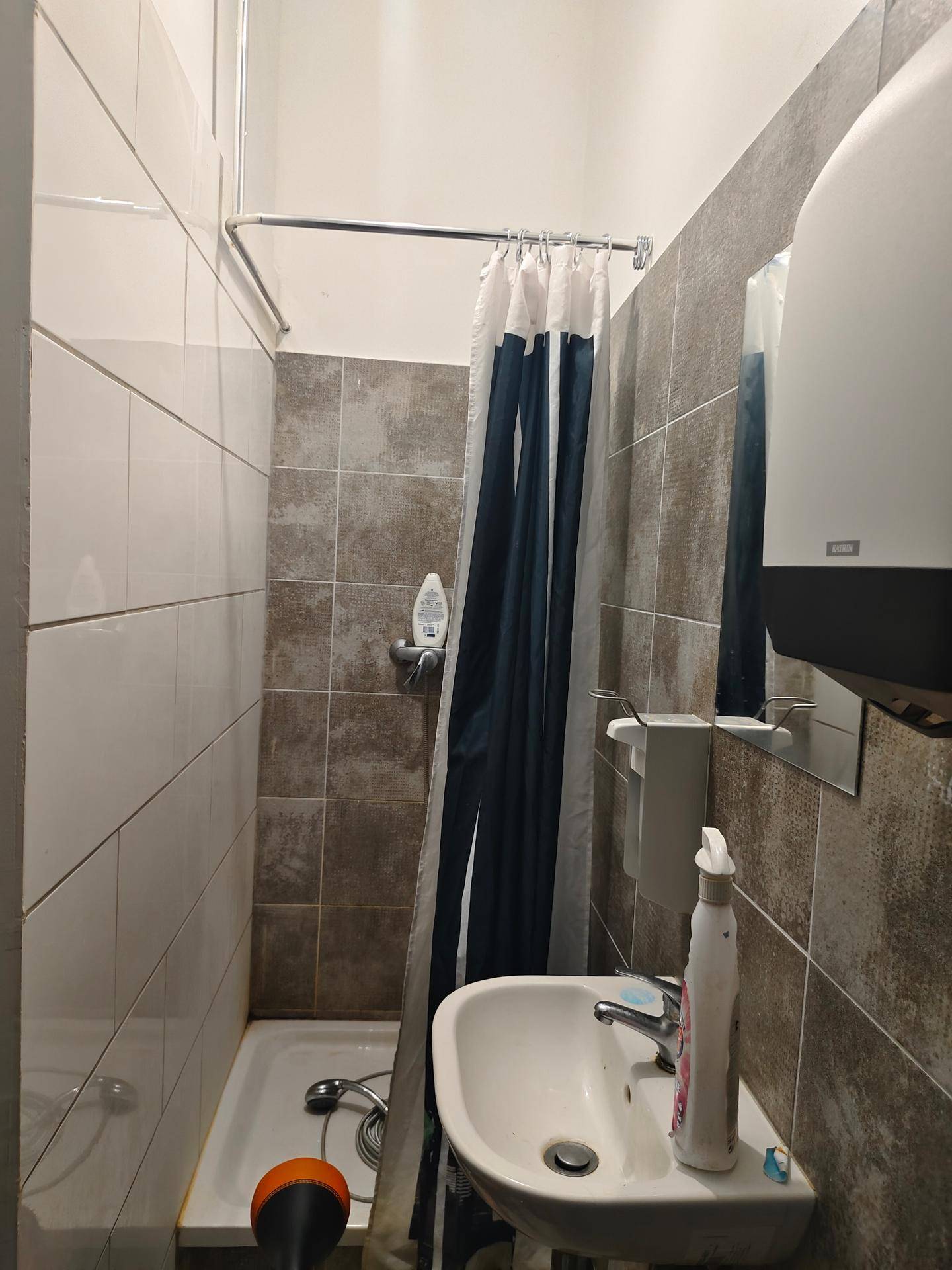 Kleines Badezimmer mit Duschwanne, Duschvorhang und Waschbecken, gefliest in Braun- und Weißtönen.