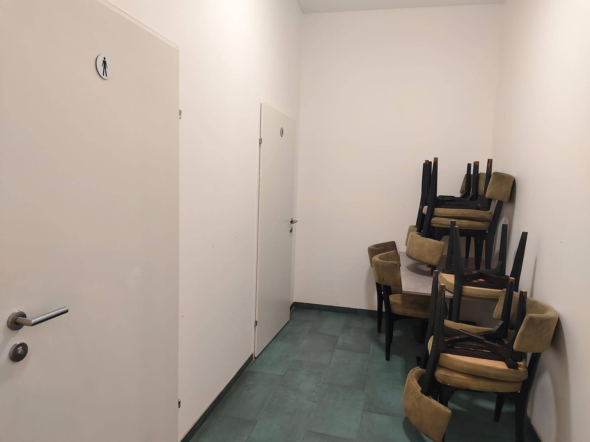 Flur mit gestapelten Stühlen und zwei Türen, eine davon mit einem Piktogramm für eine Toilette.