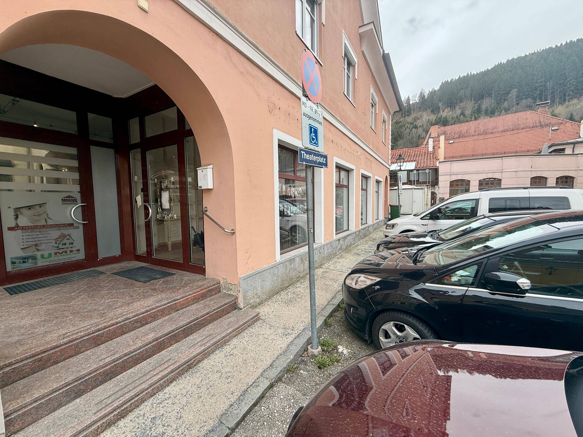 Eingangsbereich eines Gebäudes mit Rampe und Treppen, umgeben von parkenden Autos und einer Bergkulisse.
