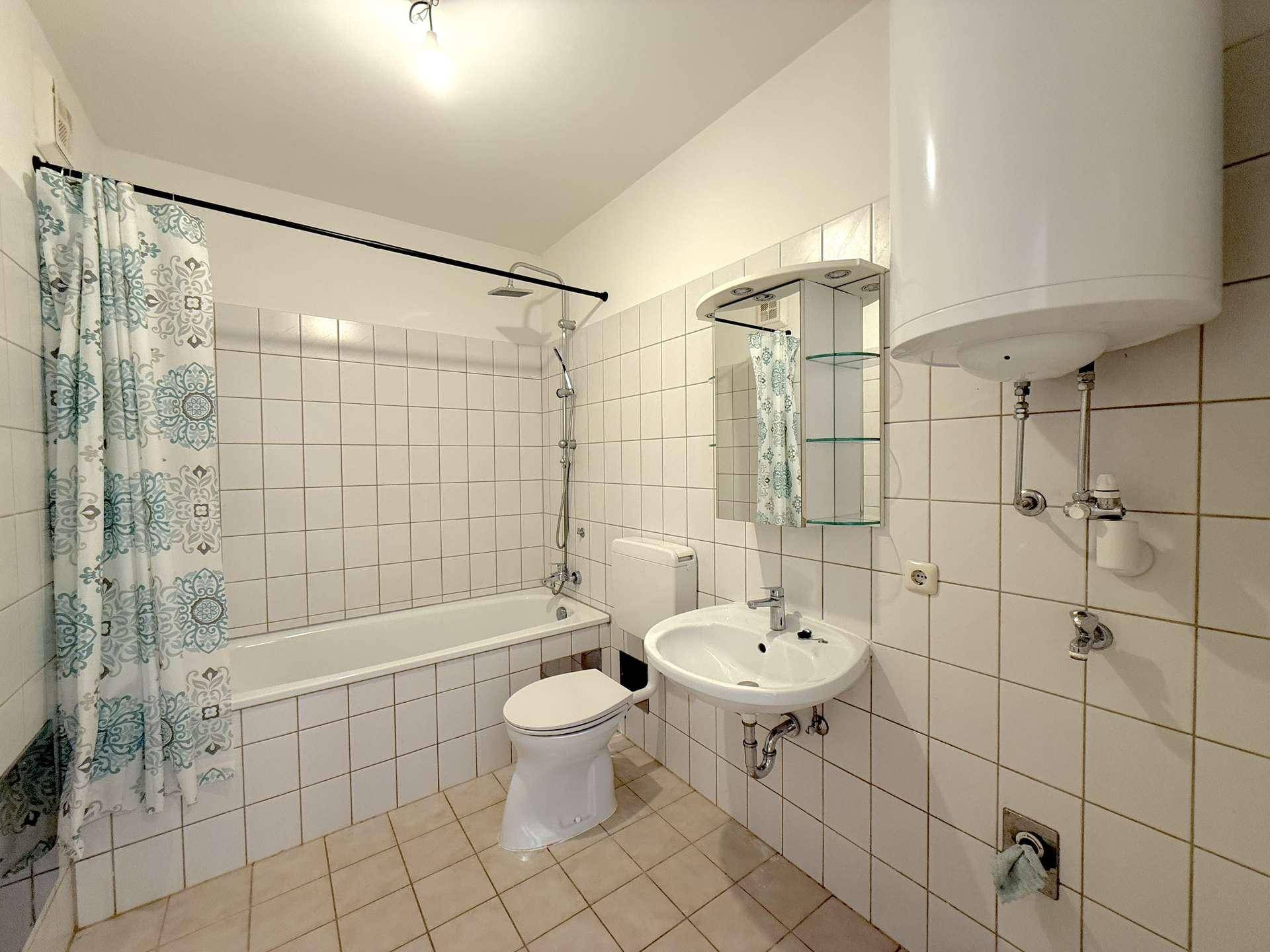 Funktionales Badezimmer mit weißen Fliesen, Badewanne mit Duschvorhang und Warmwasserboiler.