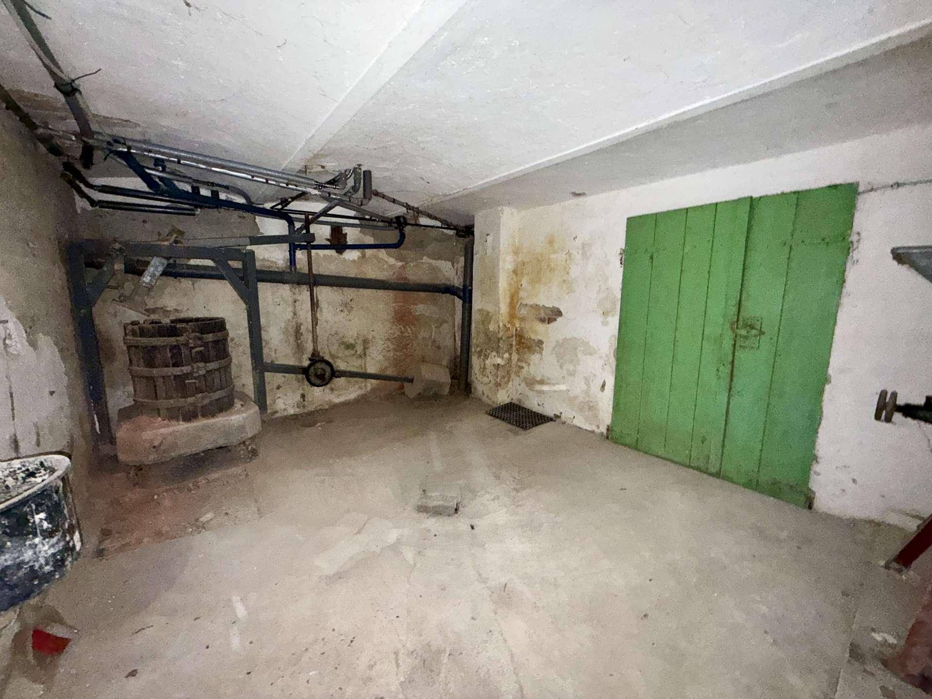 Unausgebauter Kellerraum mit Betonboden, sichtbaren Rohren und einer alten Weinpresse.