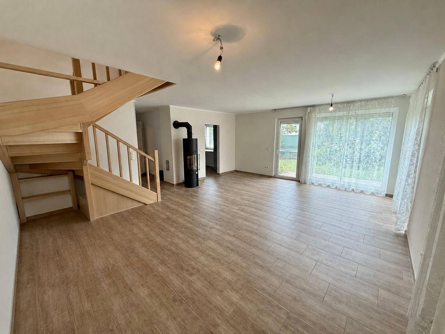 Helles Wohnzimmer mit Holztreppe, modernem Kaminofen und großen Fenstern zum Garten.