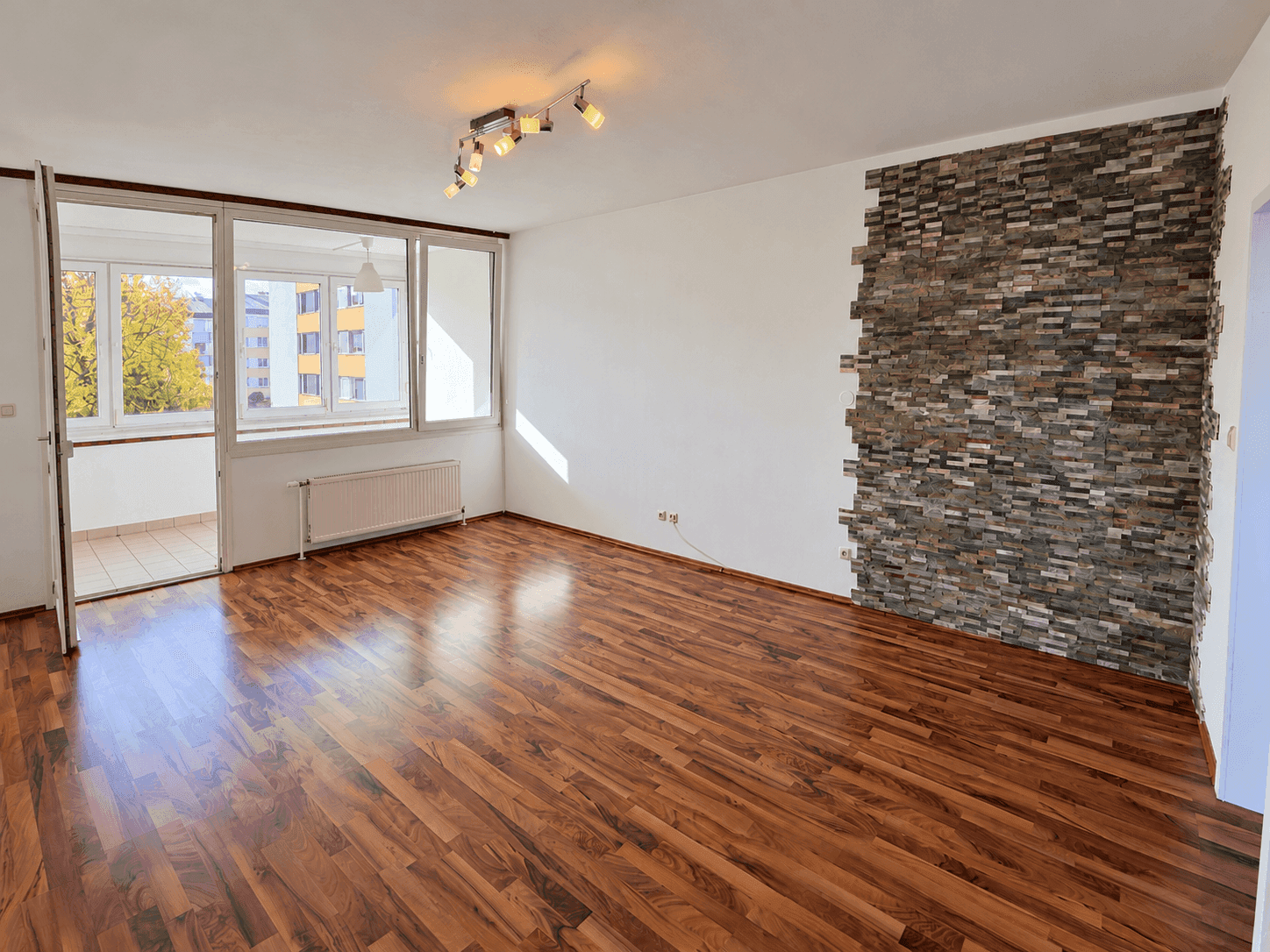 Geräumiges Wohnzimmer mit Laminatboden, dekorativer Steinwand und direktem Zugang zum Balkon.