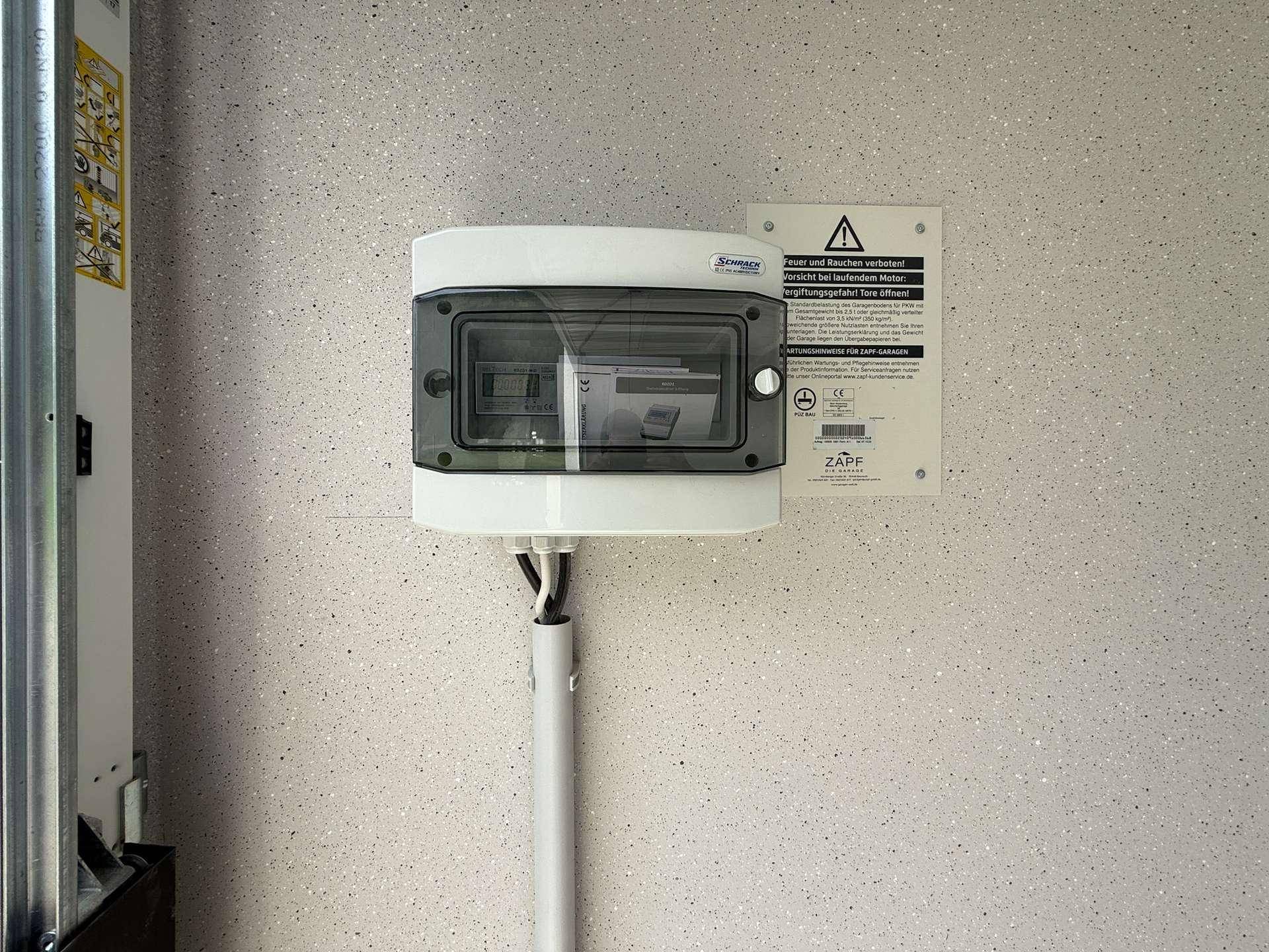 Nahaufnahme eines elektrischen Schaltkastens und eines Warnschildes an der strukturierten Garagenwand.