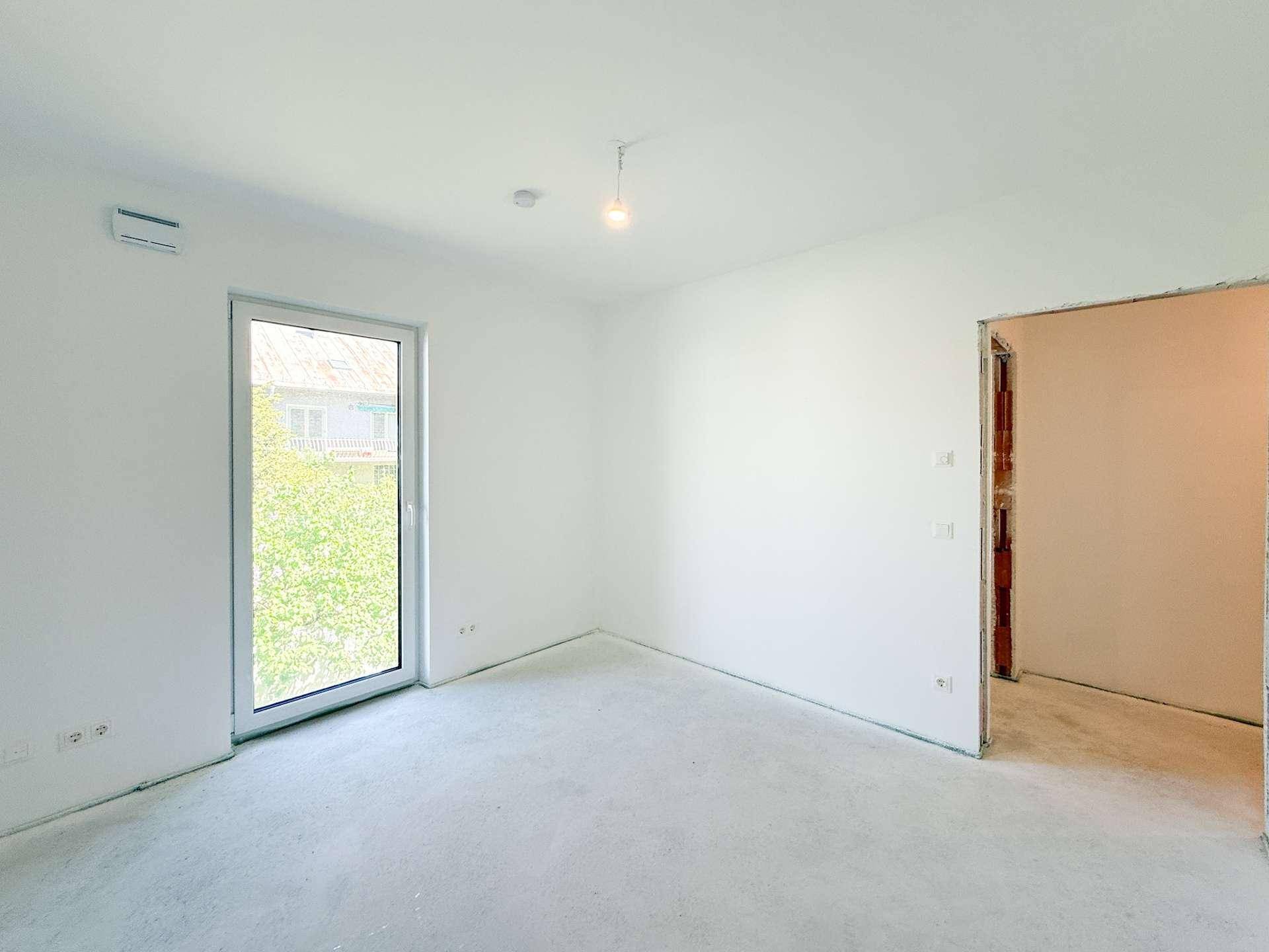 Helles, unmöbliertes Zimmer im Rohbau mit Fenster und Zugang zum Außenbereich.