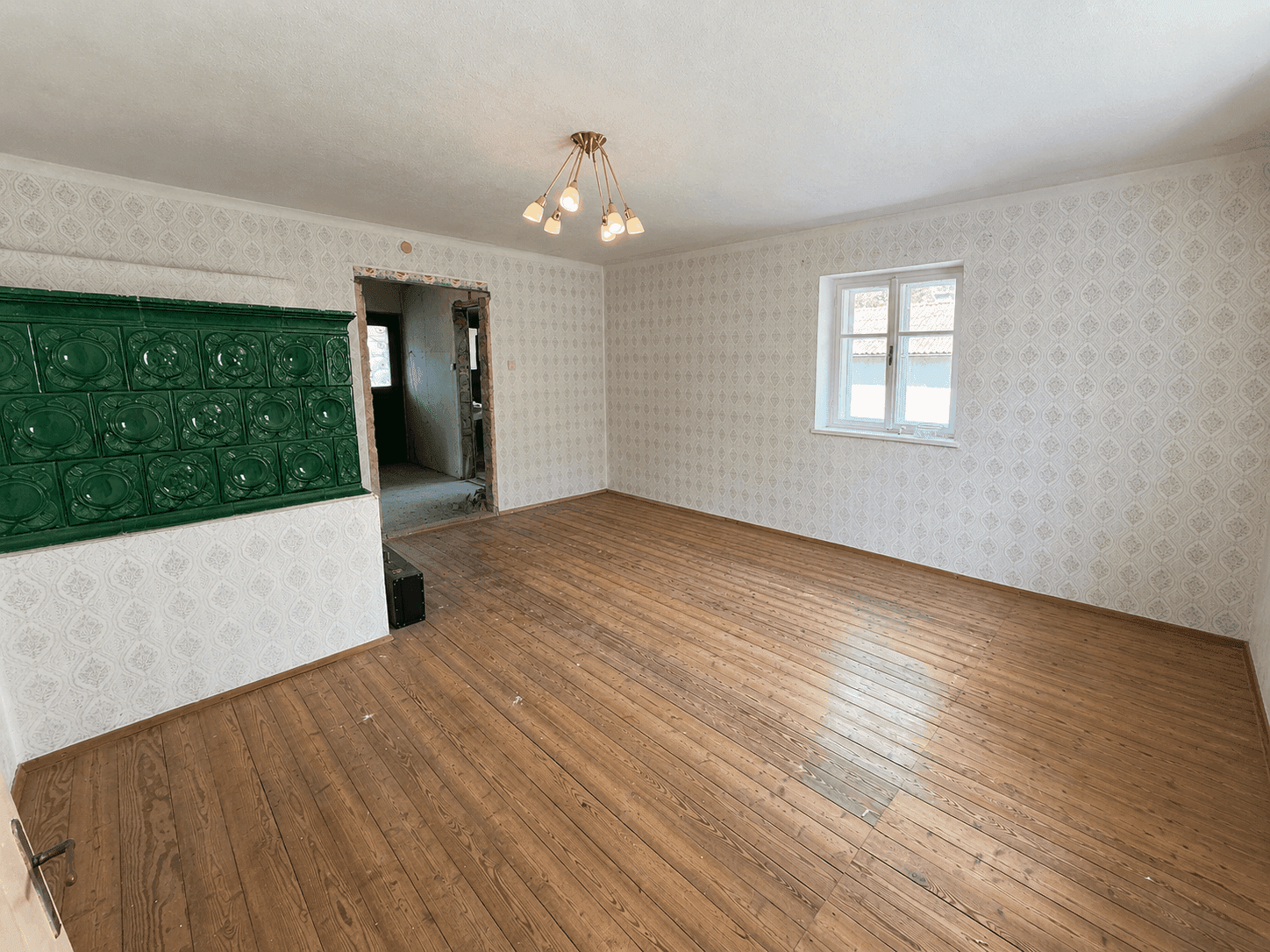 Wohnzimmer mit traditionellem Kachelofen, Holzboden und alter Tapete, bereit zur Renovierung.