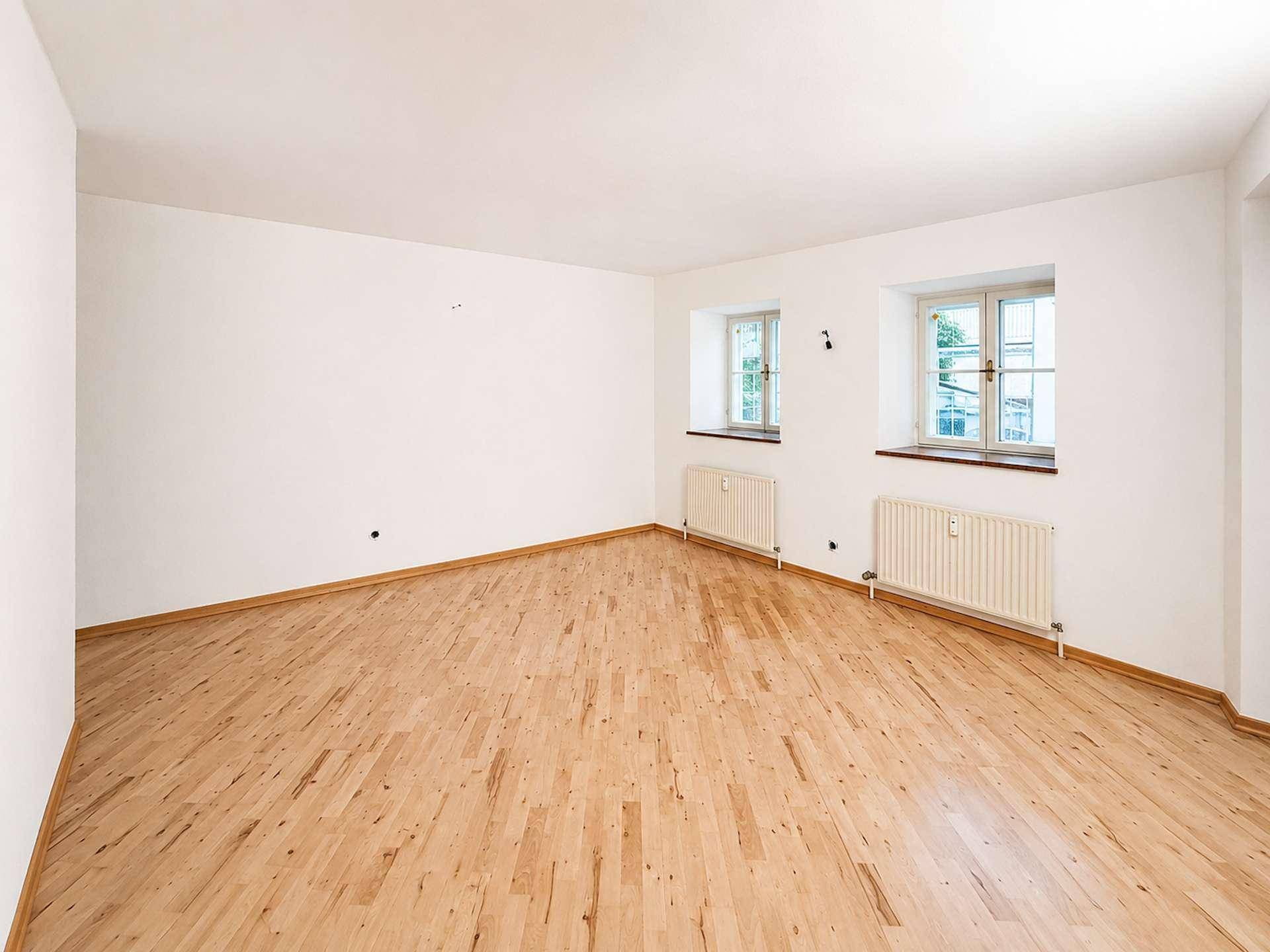 Leerer Wohnraum mit Holzboden und zwei Fenstern, bereit für neue Bewohner und deren Gestaltungsideen.