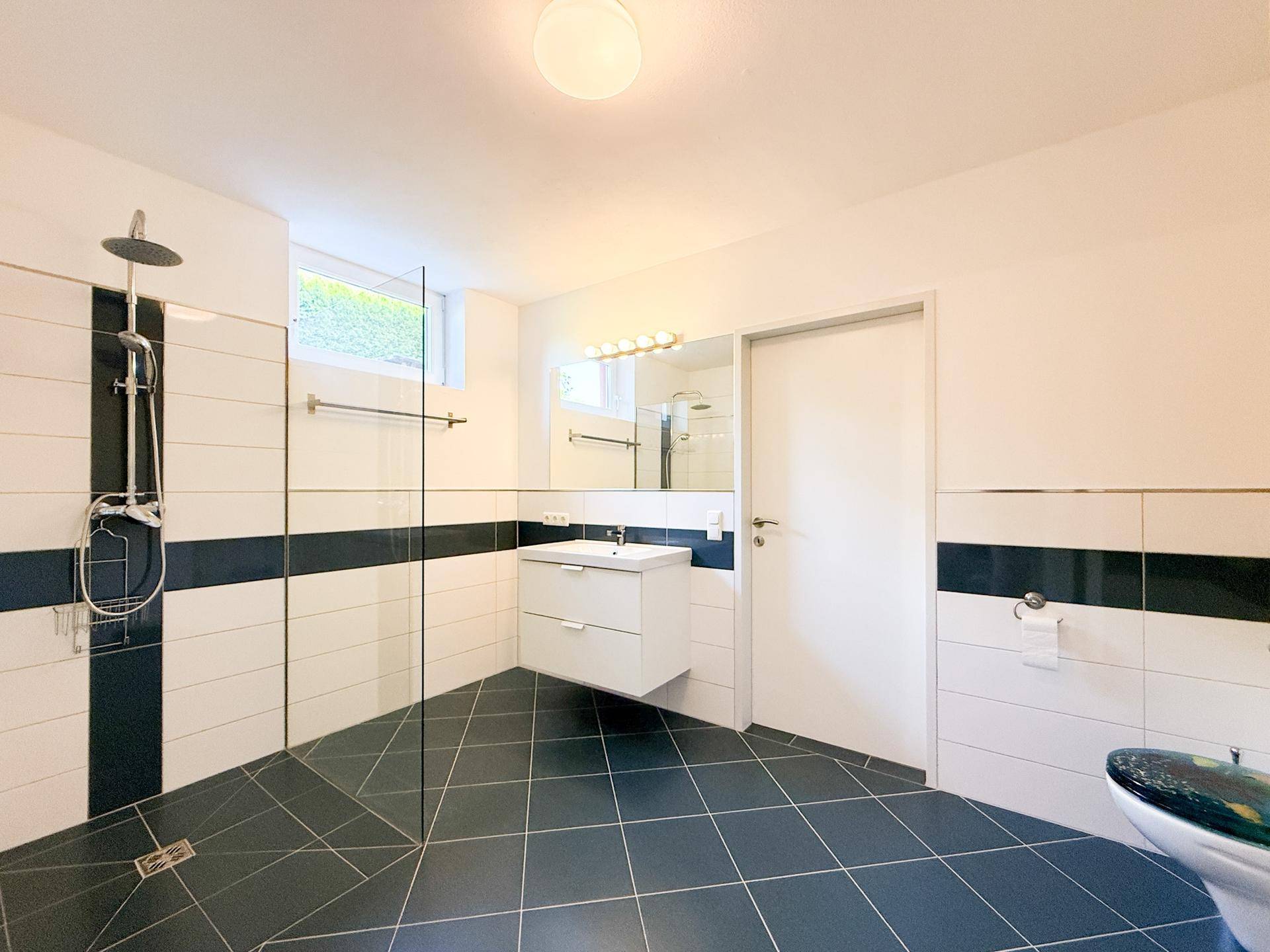 Modernes Badezimmer mit ebenerdiger Dusche, Waschbecken und Fenster, in Weiß und Dunkelblau gefliest.