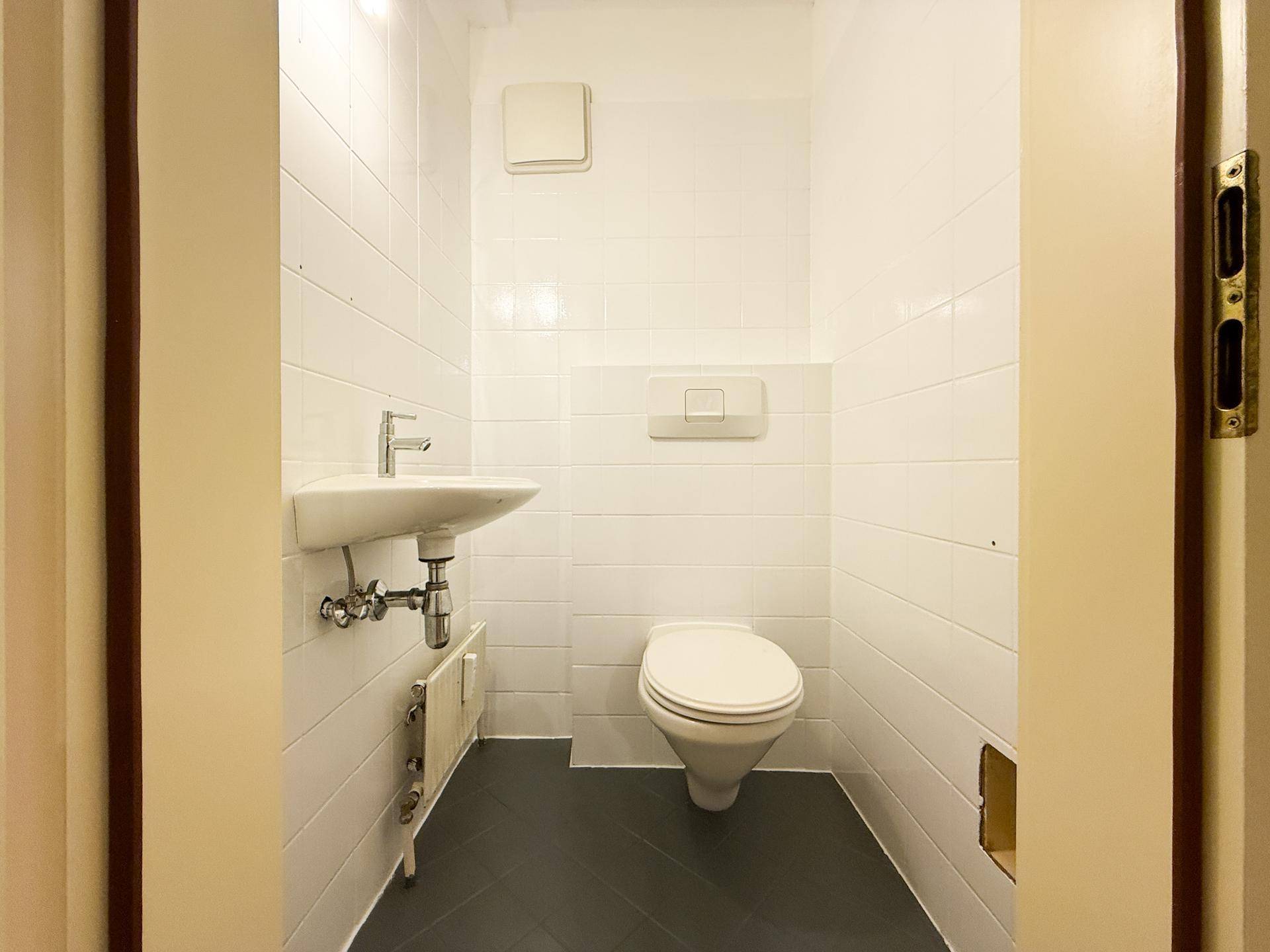 Separates WC mit Wand-WC, kleinem Waschbecken und weißen Fliesen.