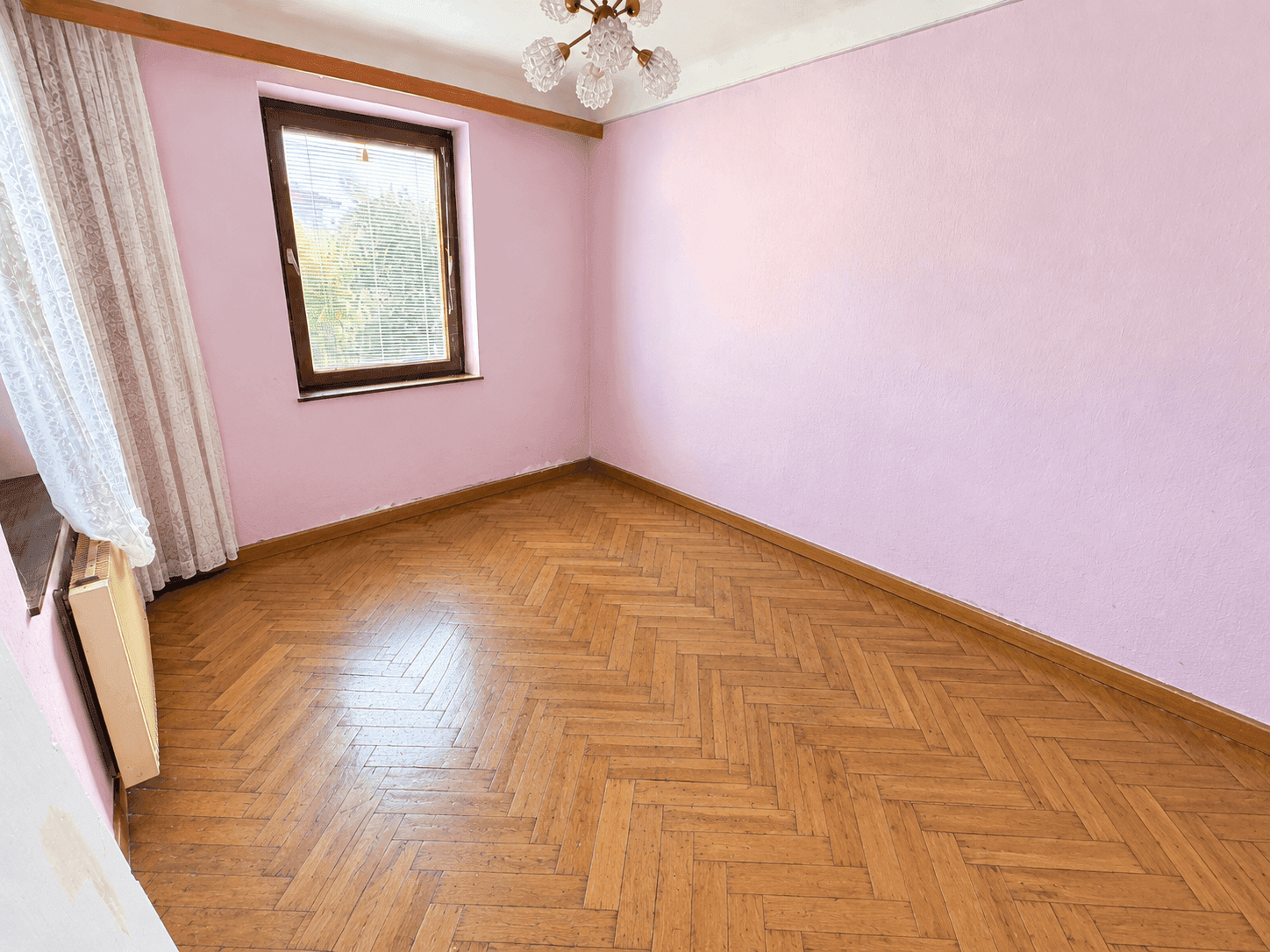 Leeres Zimmer mit rosa Wänden, Fischgrätparkett und einem Fenster, das Tageslicht spendet.