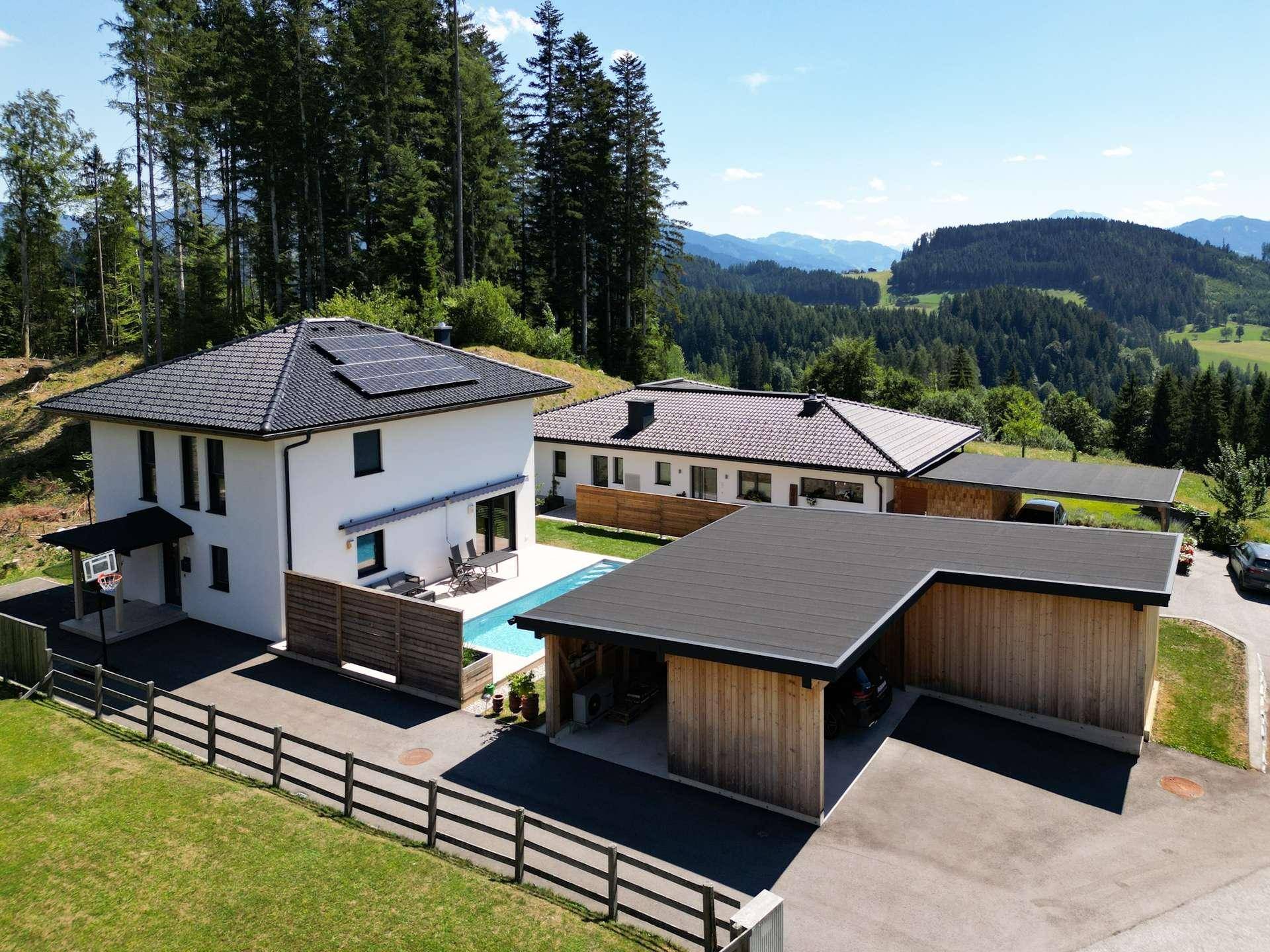 Luftaufnahme eines Anwesens mit zwei Häusern, Pool und Carport, eingebettet in eine grüne Berglandschaft.