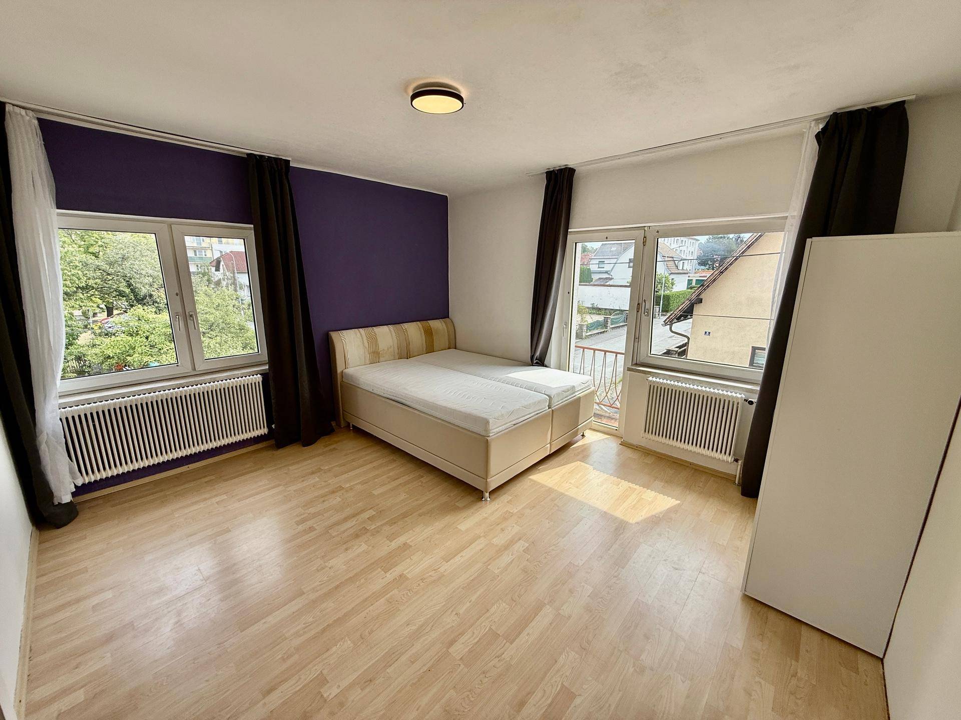 Helles Schlafzimmer mit Holzboden, lila Akzentwand und Zugang zu einem französischen Balkon.