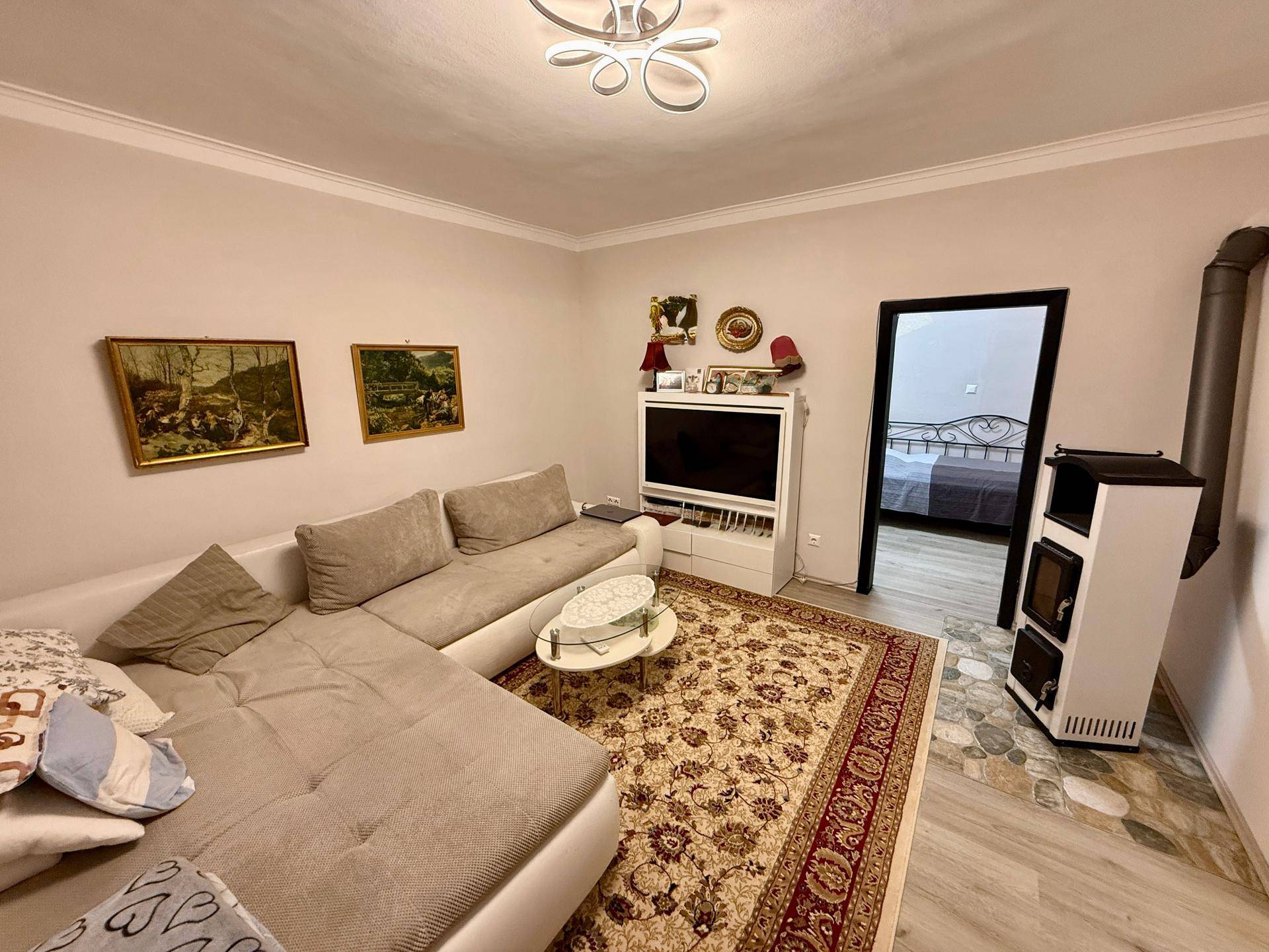 Gemütliches Wohnzimmer mit Ecksofa, Teppich und einem modernen Holzofen, der zum Schlafzimmer führt.