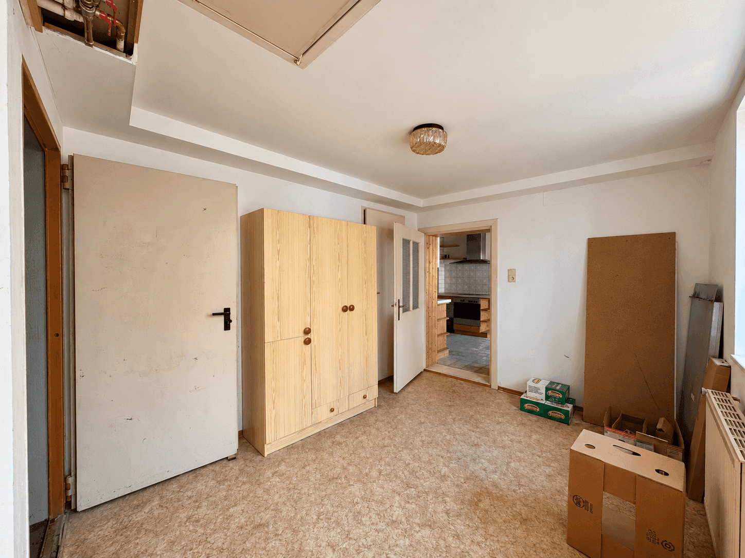 Einfaches Zimmer mit Teppichboden, Kleiderschrank und Blick in die Küche.