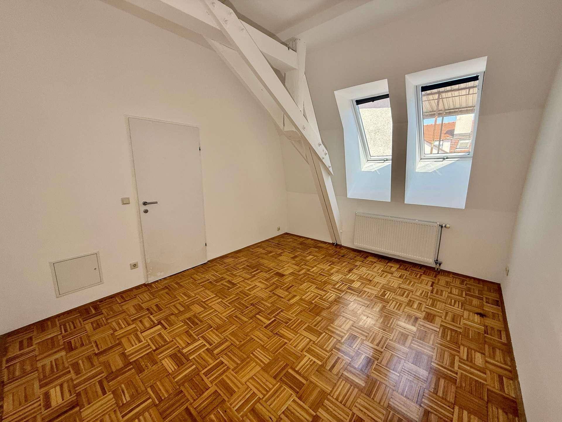 Geräumiges Dachzimmer mit sichtbaren Holzbalken, Parkettboden und zwei Dachfenstern für natürliche Belichtung.