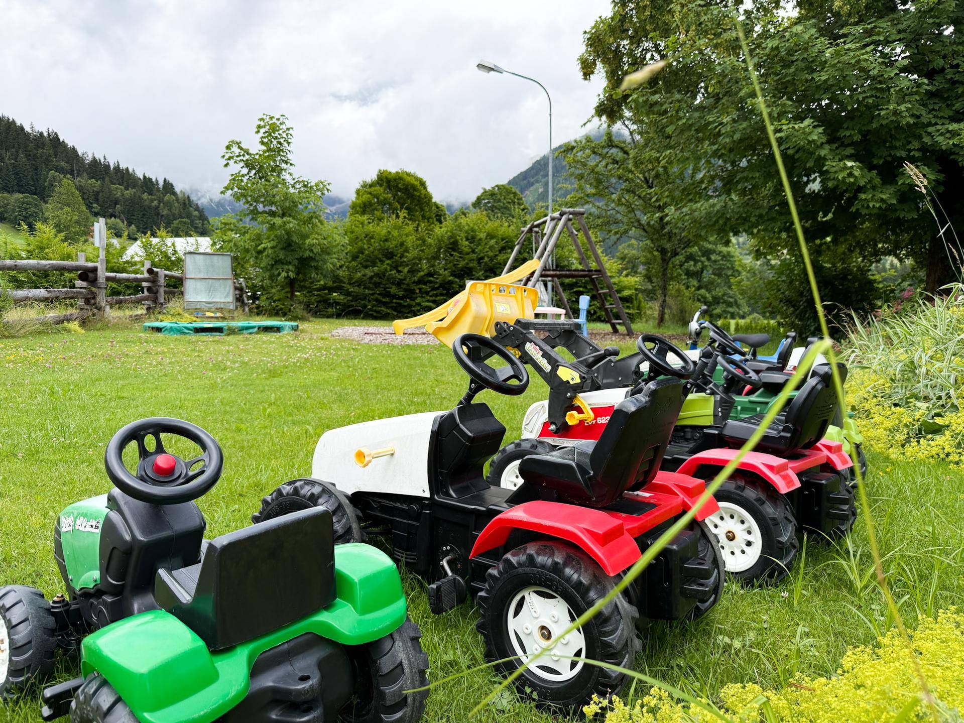 Spielbereich im Freien mit mehreren Kindertraktoren auf einer grünen Wiese, umgeben von Bäumen und Bergen im Hintergrund.